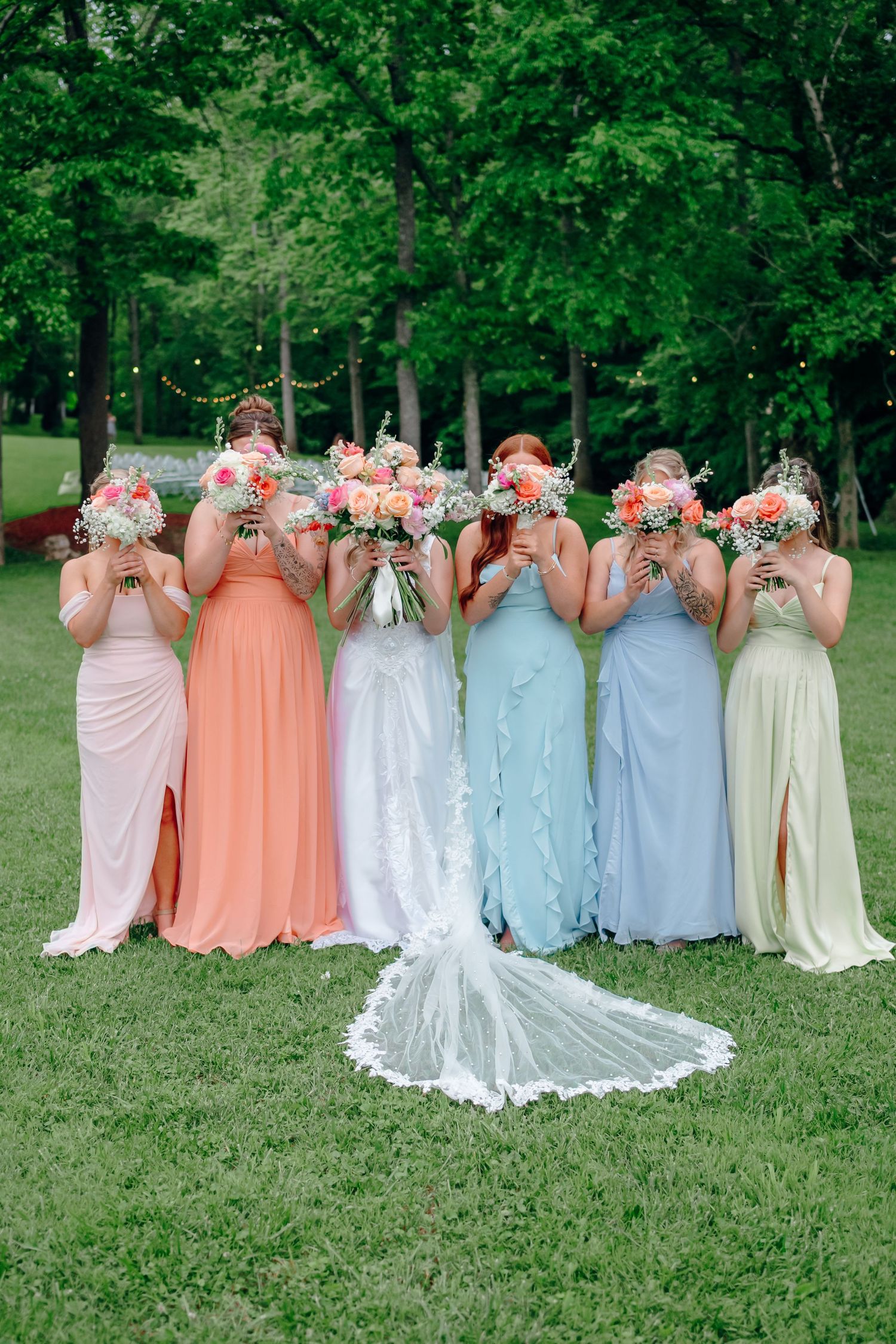 Bridesmaids in pastel colored dresses lined up holding bouquets against their faces.