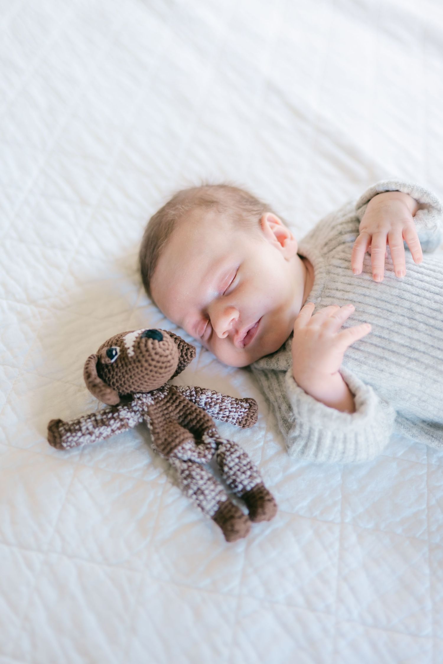 Baby sleeping peacefully on white bedding with small brown knitted stuffed animal toy.