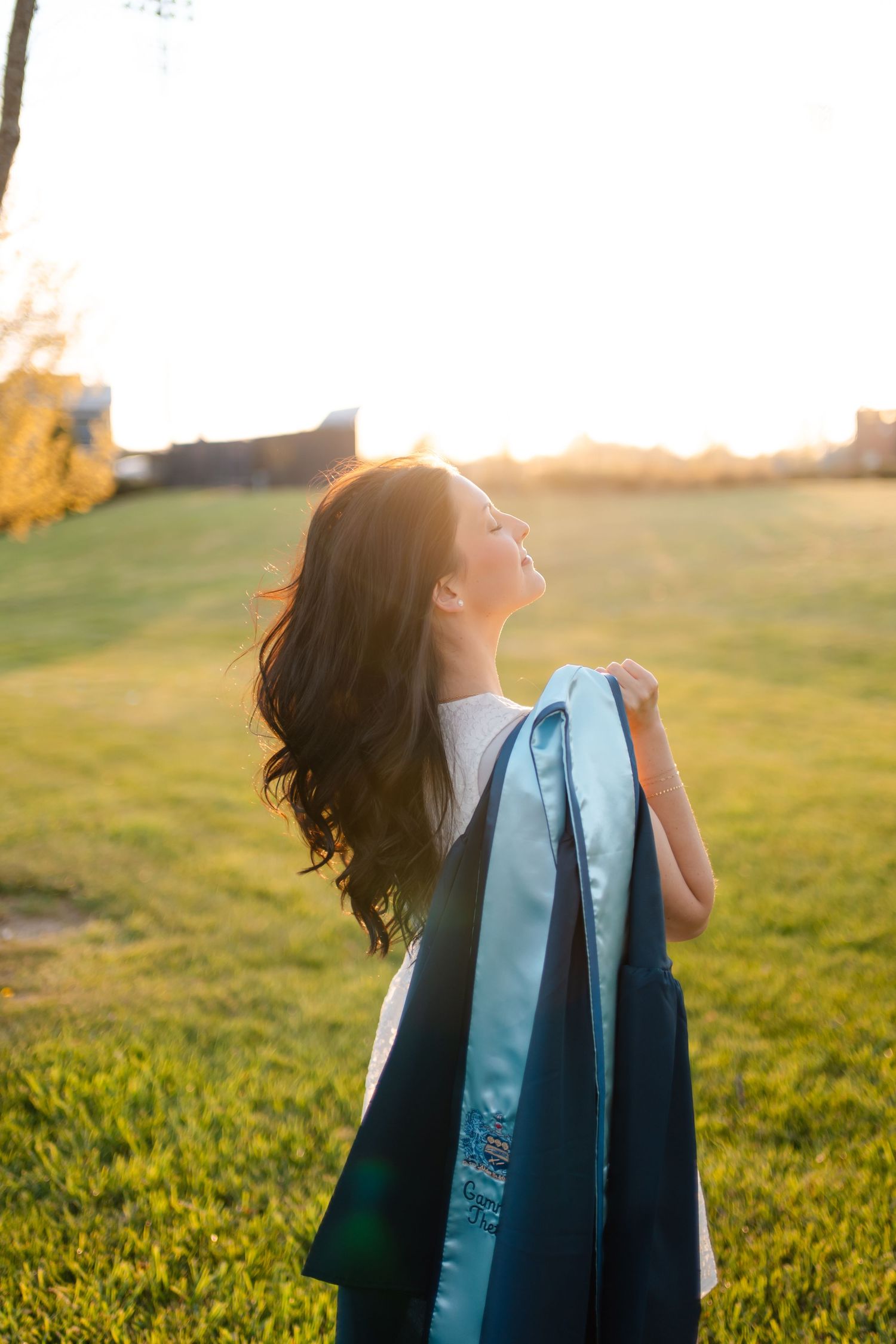 Young woman in graduation gown holding her cap, standing in sunlit green field, celebrating her academic achievement.