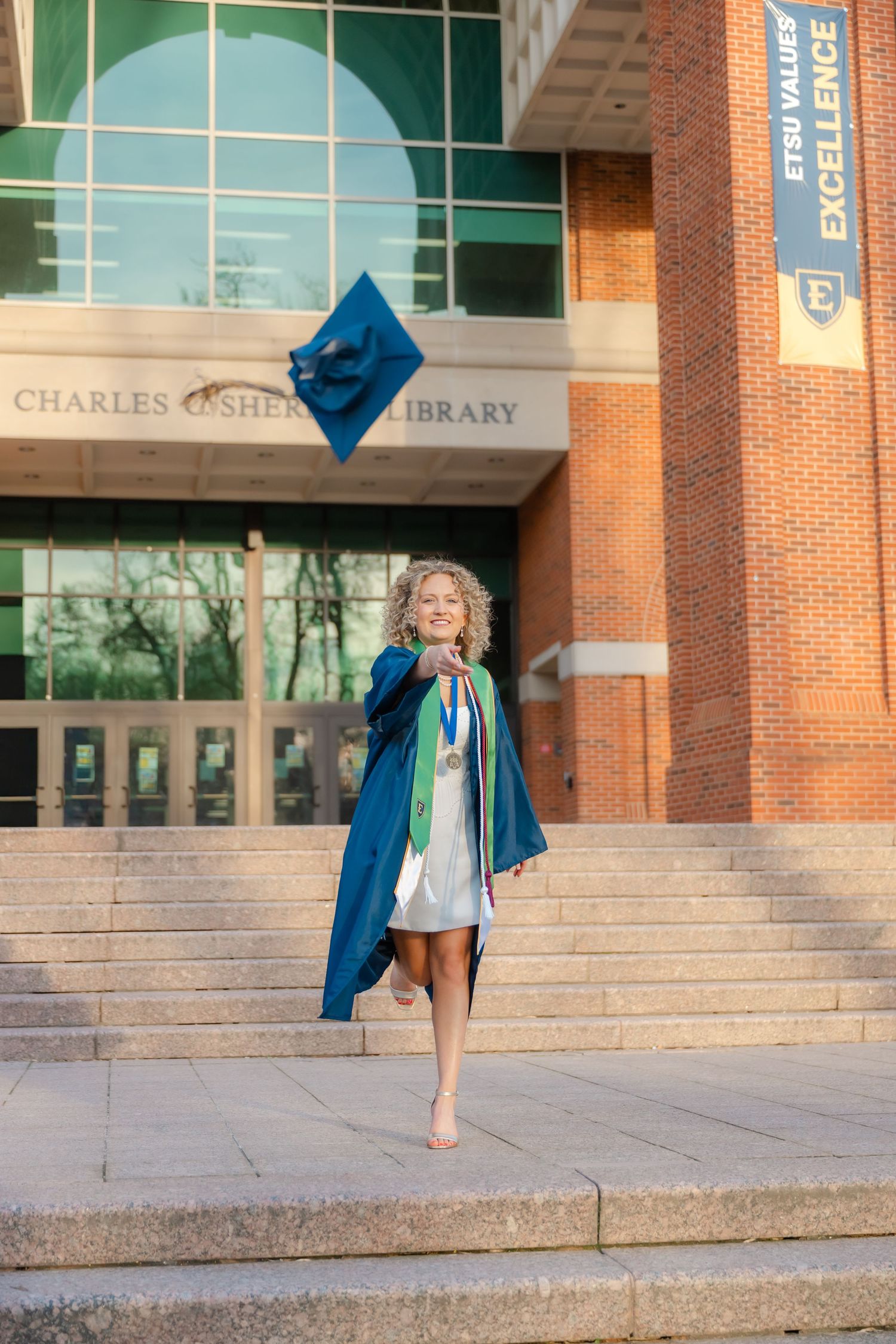 A joyful graduate in a blue cap and gown tosses her cap outside Charles Sherrod Library at ETSU.