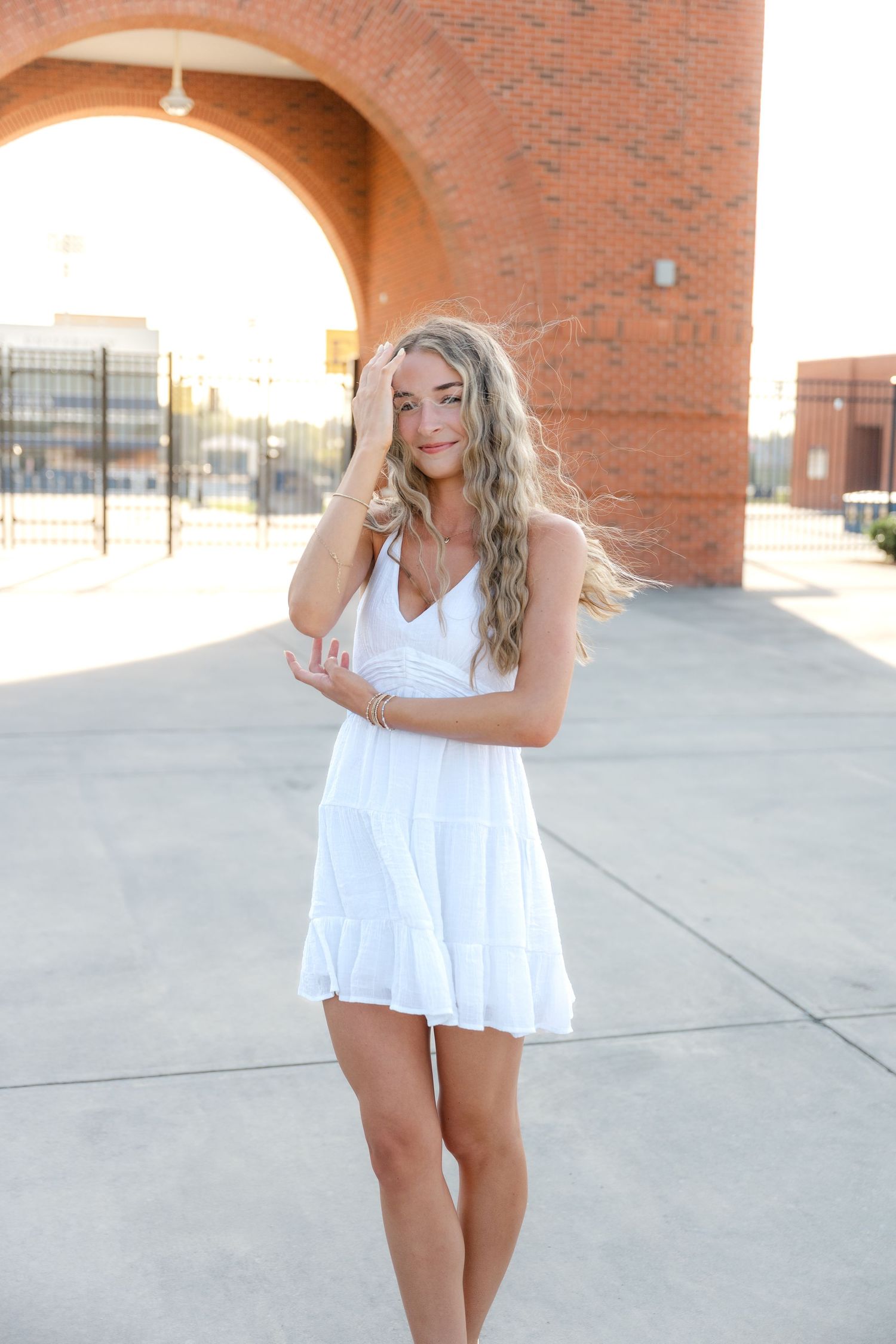 Young woman in white sundress poses outdoors near brick archway, long blonde wavy hair blowing in breeze.