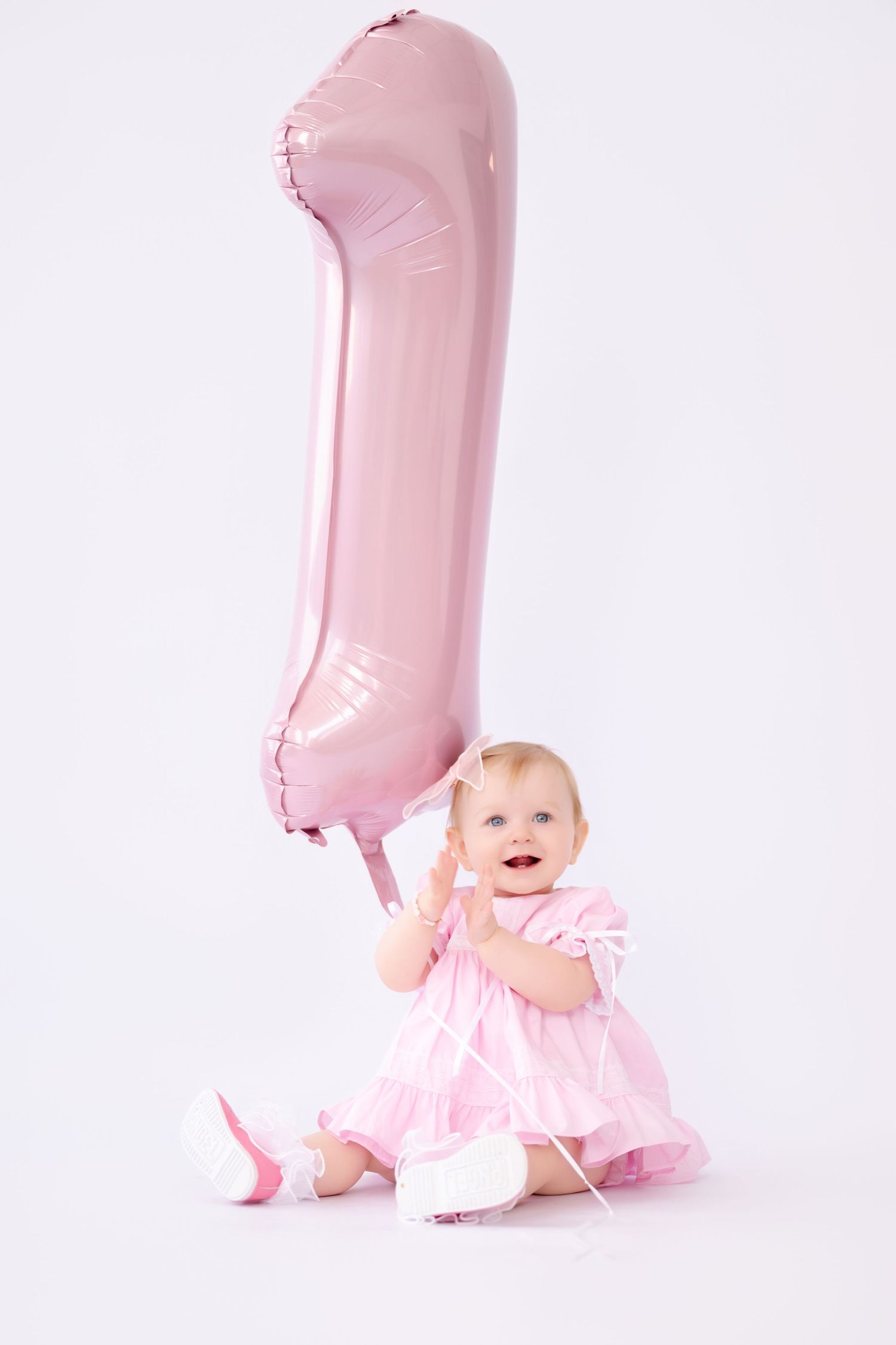 Baby girl in pink dress sitting on white background holding a large pink number 1 balloon for her first birthday.