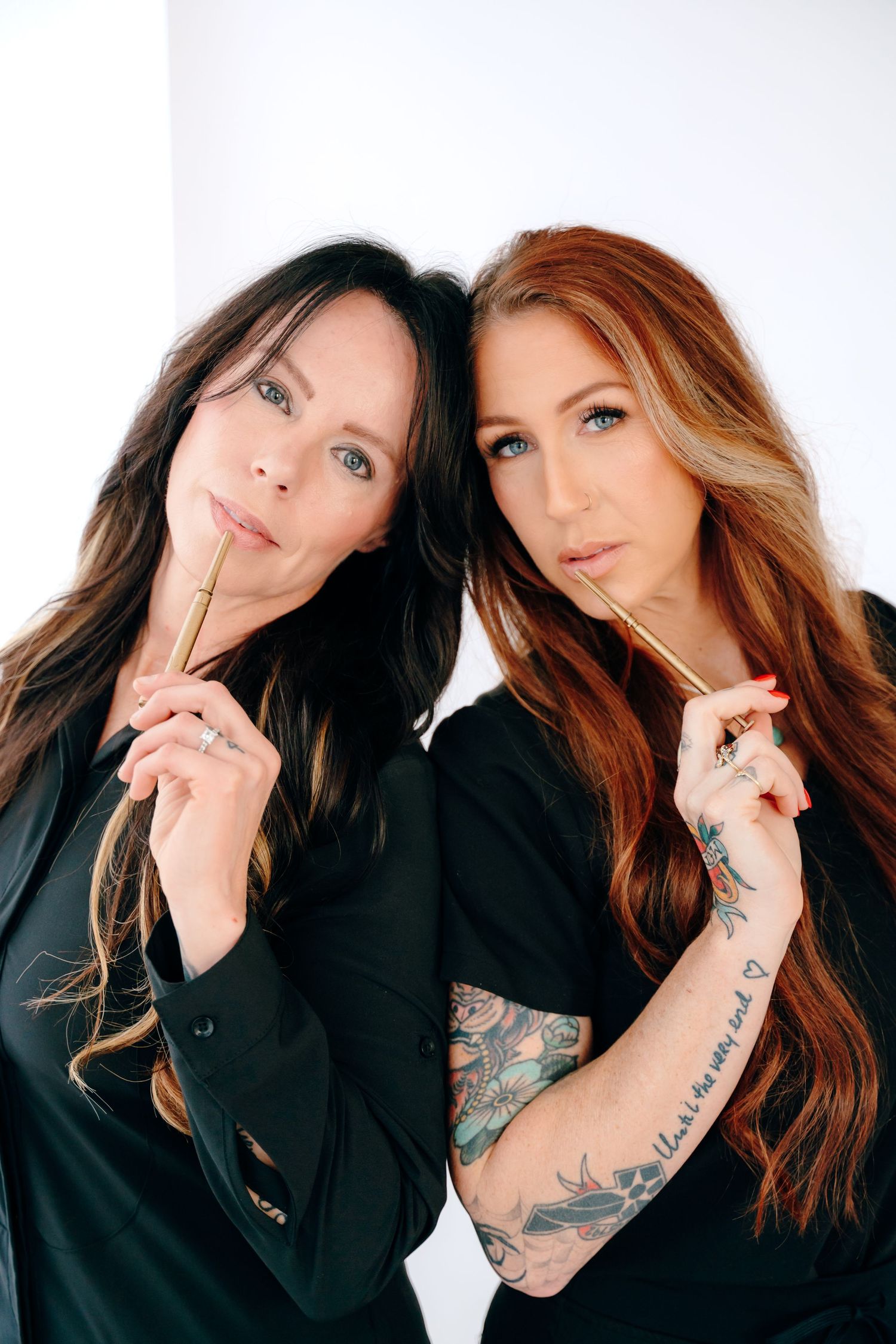 Two women with long hair in black outfits pose together holding pens against a white backdrop.