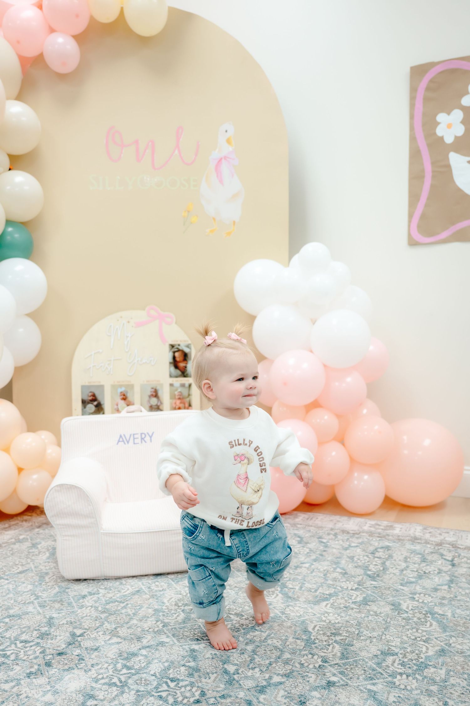 A baby in blue jeans crawls on a floral rug at a party decorated with pink and white balloons.