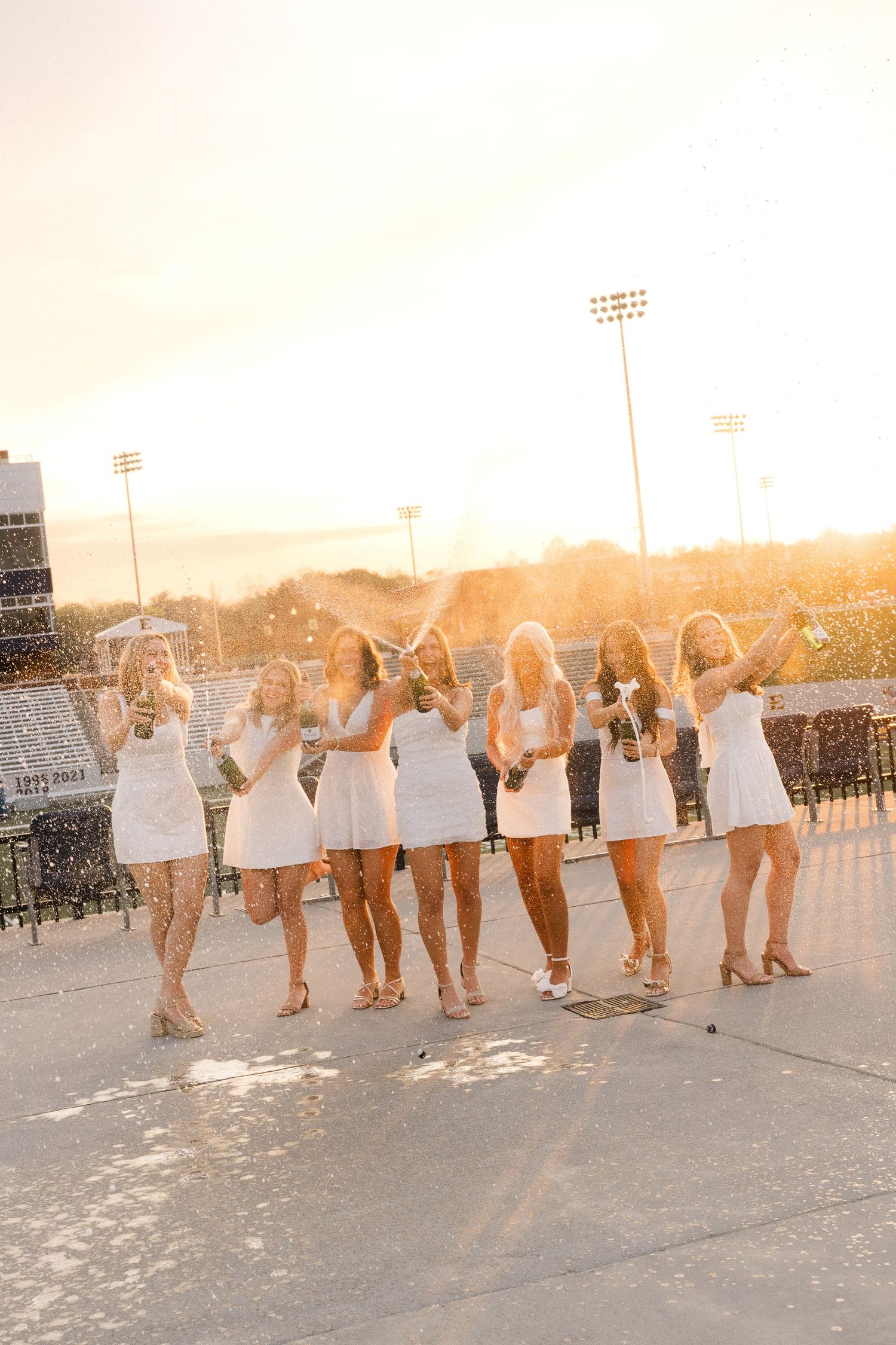 Seven women in white dresses celebrate with champagne at a stadium during golden hour sunset.