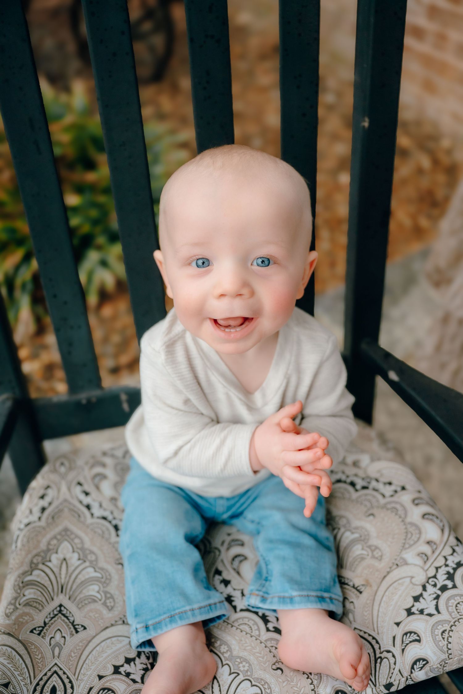 Baby wearing white top and blue pants sits on a decorative cushion against black railings while smiling.