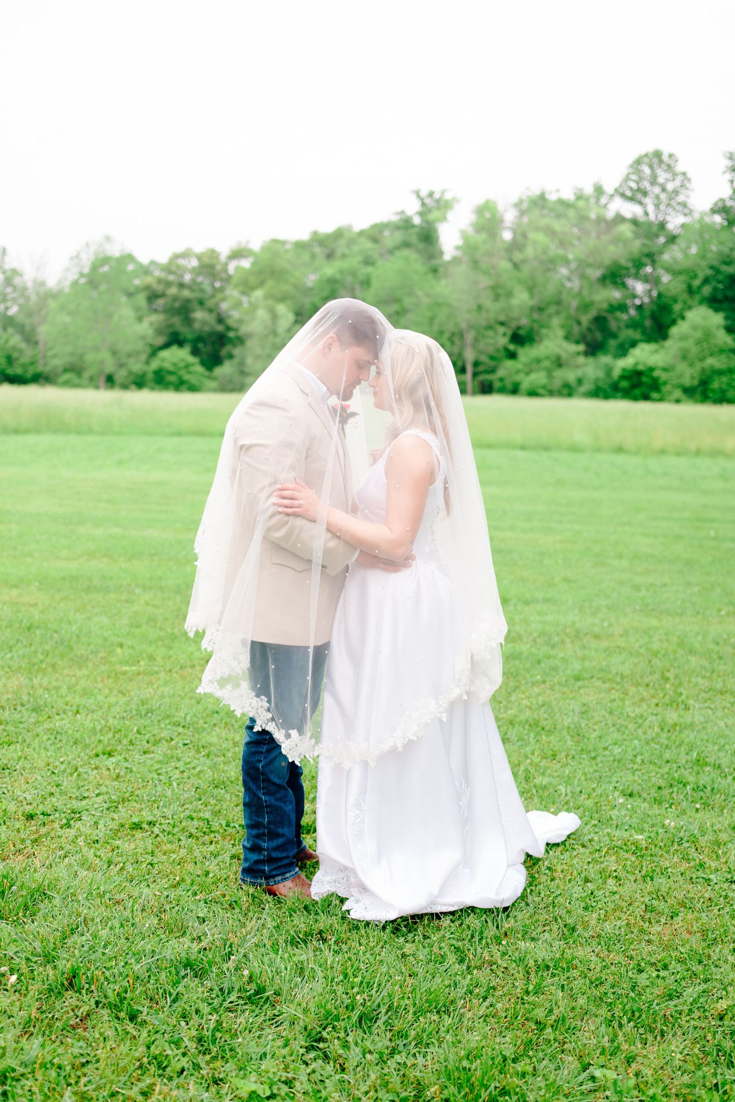 Wedding couple poses on green lawn with flowing white veil creating ethereal effect.