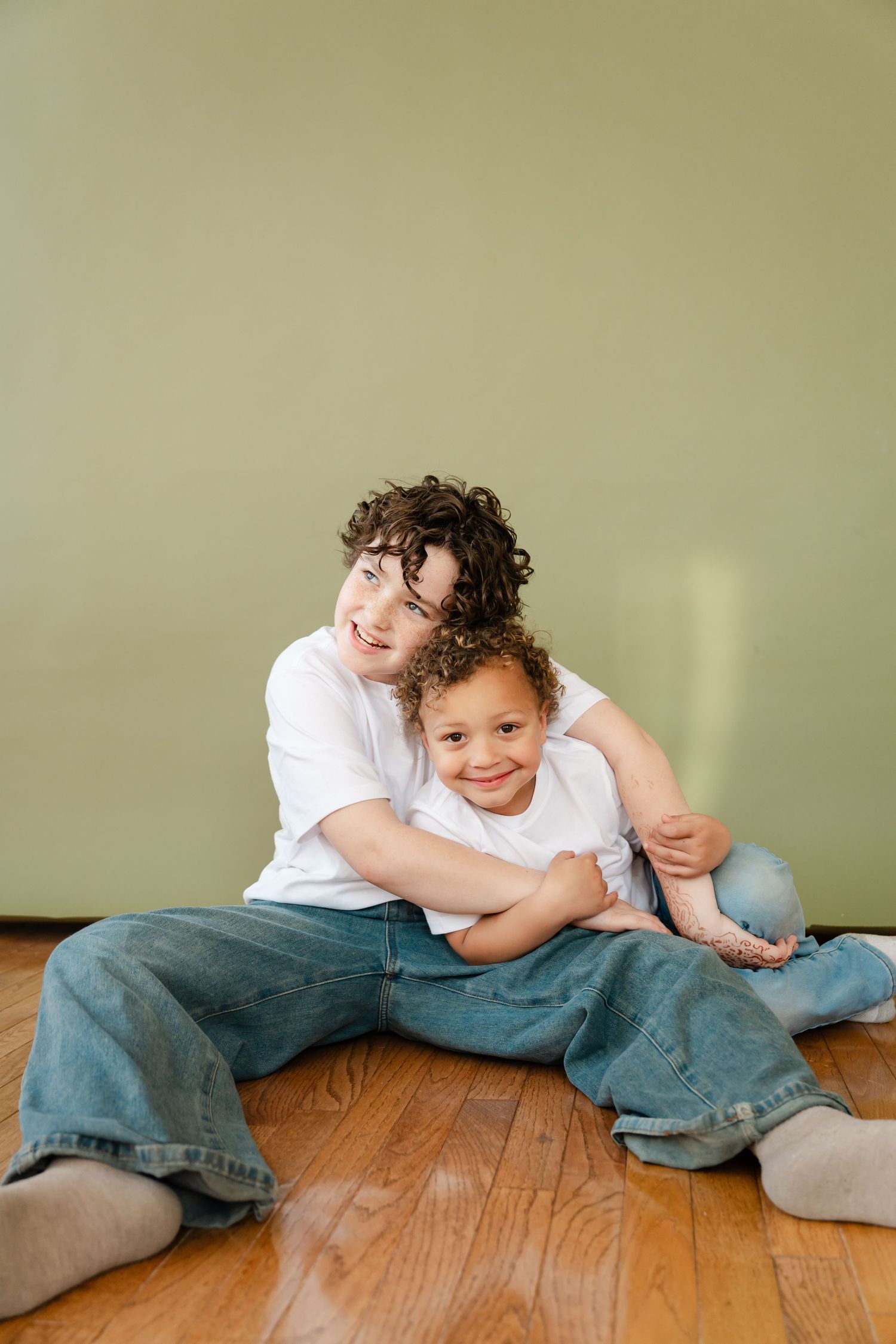 Two children sitting on hardwood floor against green wall, older child hugging younger one, both wearing white tees and jeans.