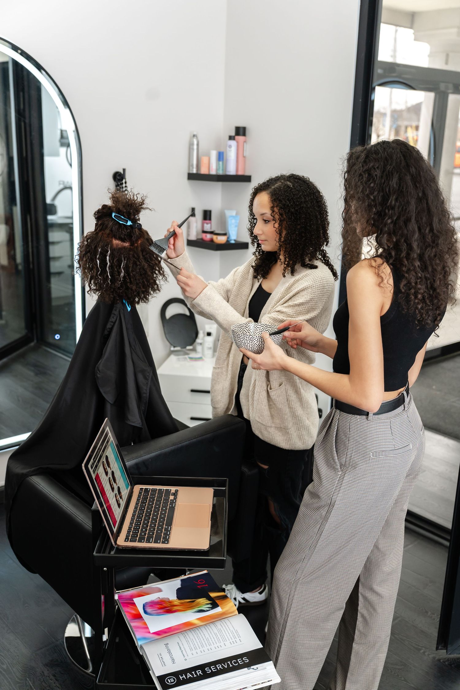Stylists working on curly hair while referencing a laptop in a modern salon setting.