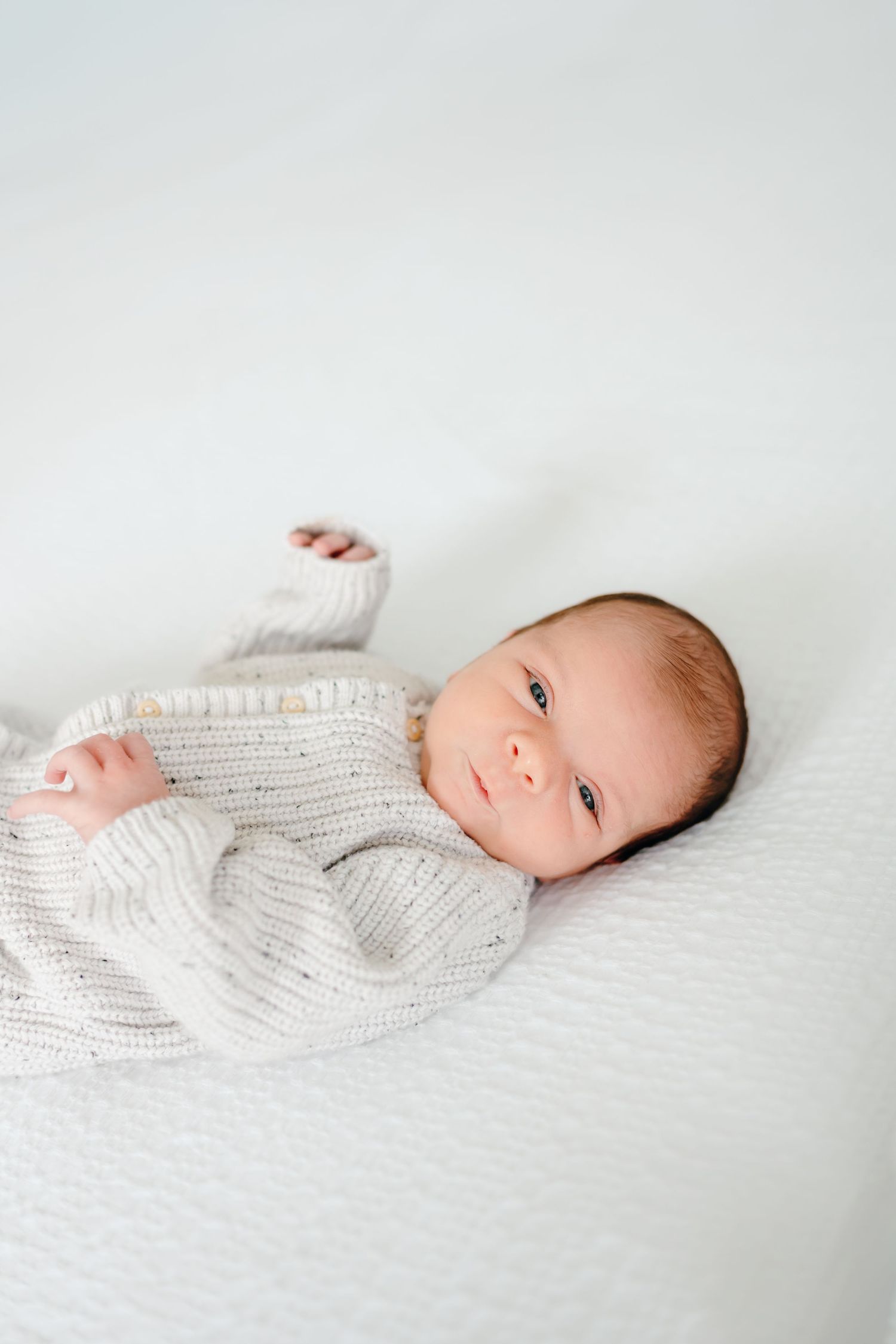 Newborn baby wearing white knit sweater lying on textured white blanket.