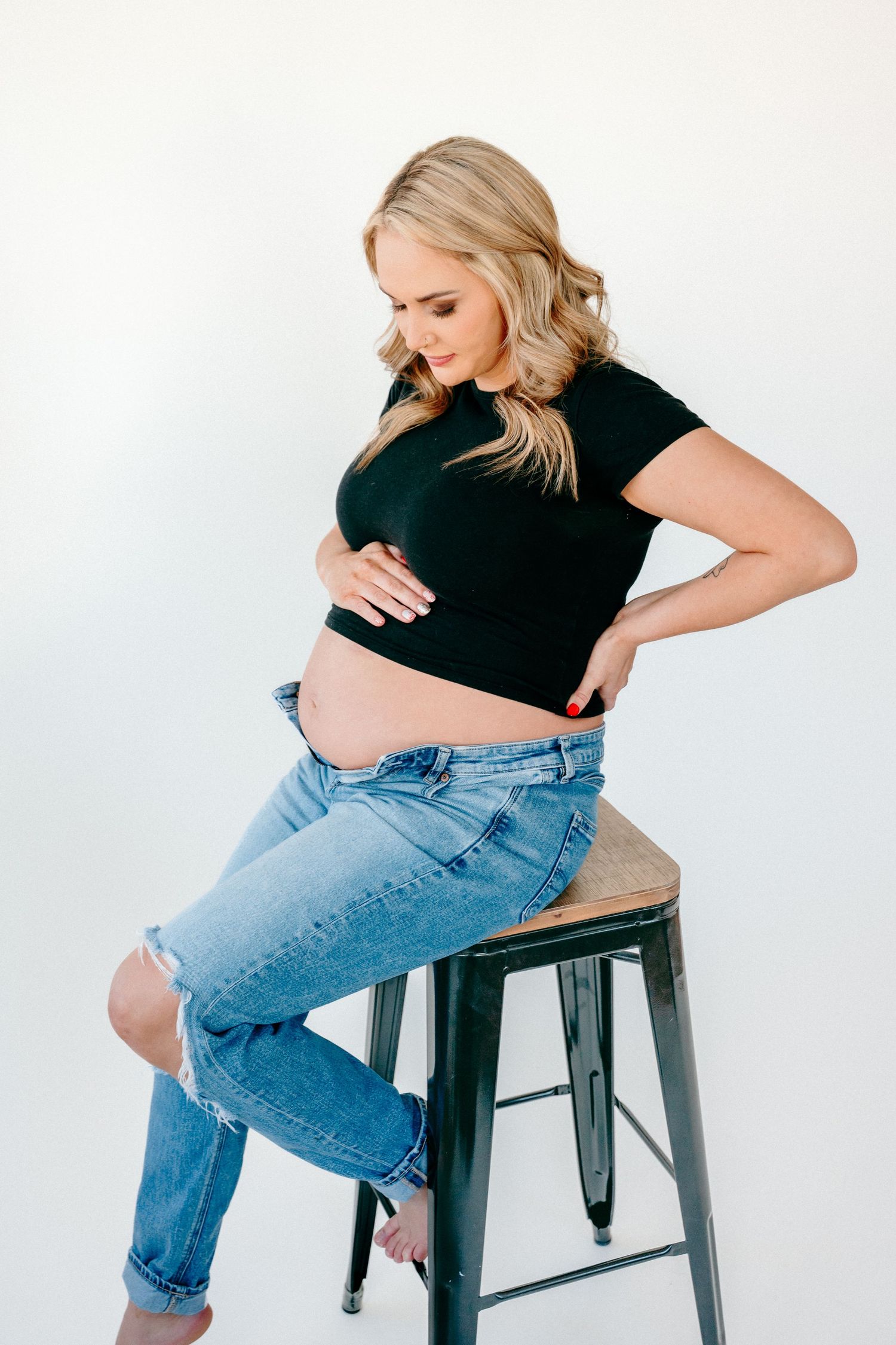 Maternity photo series showing a woman in jeans and black top sitting on an industrial stool.