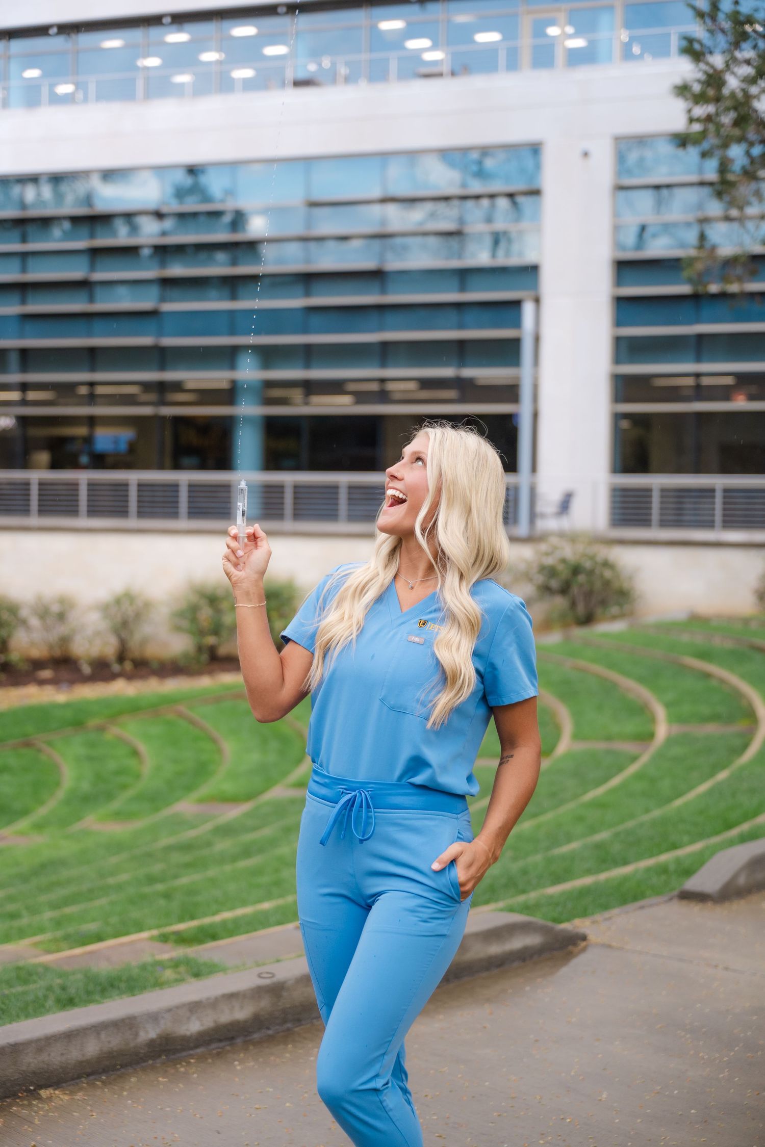 Smiling blonde woman in blue scrubs holds a pen outdoors near a modern glass building with green landscaping.