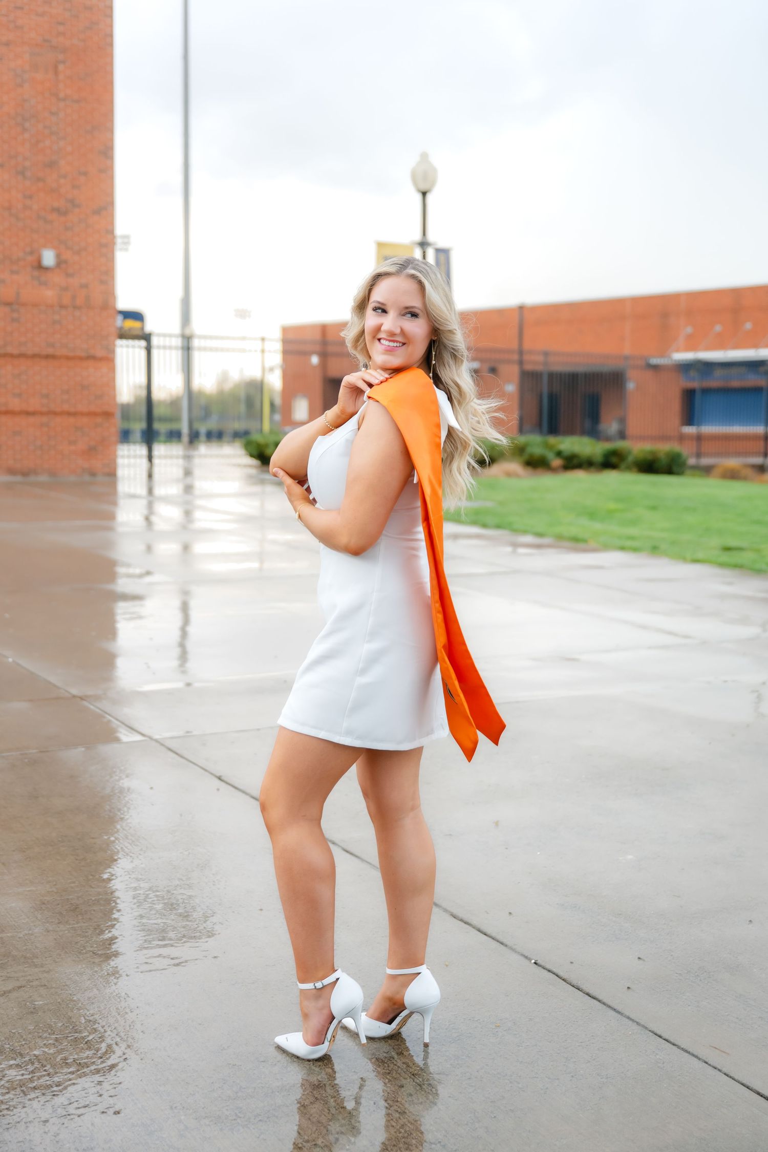 Smiling blonde woman in white mini dress and heels with orange sash poses outdoors on wet pavement near brick building.