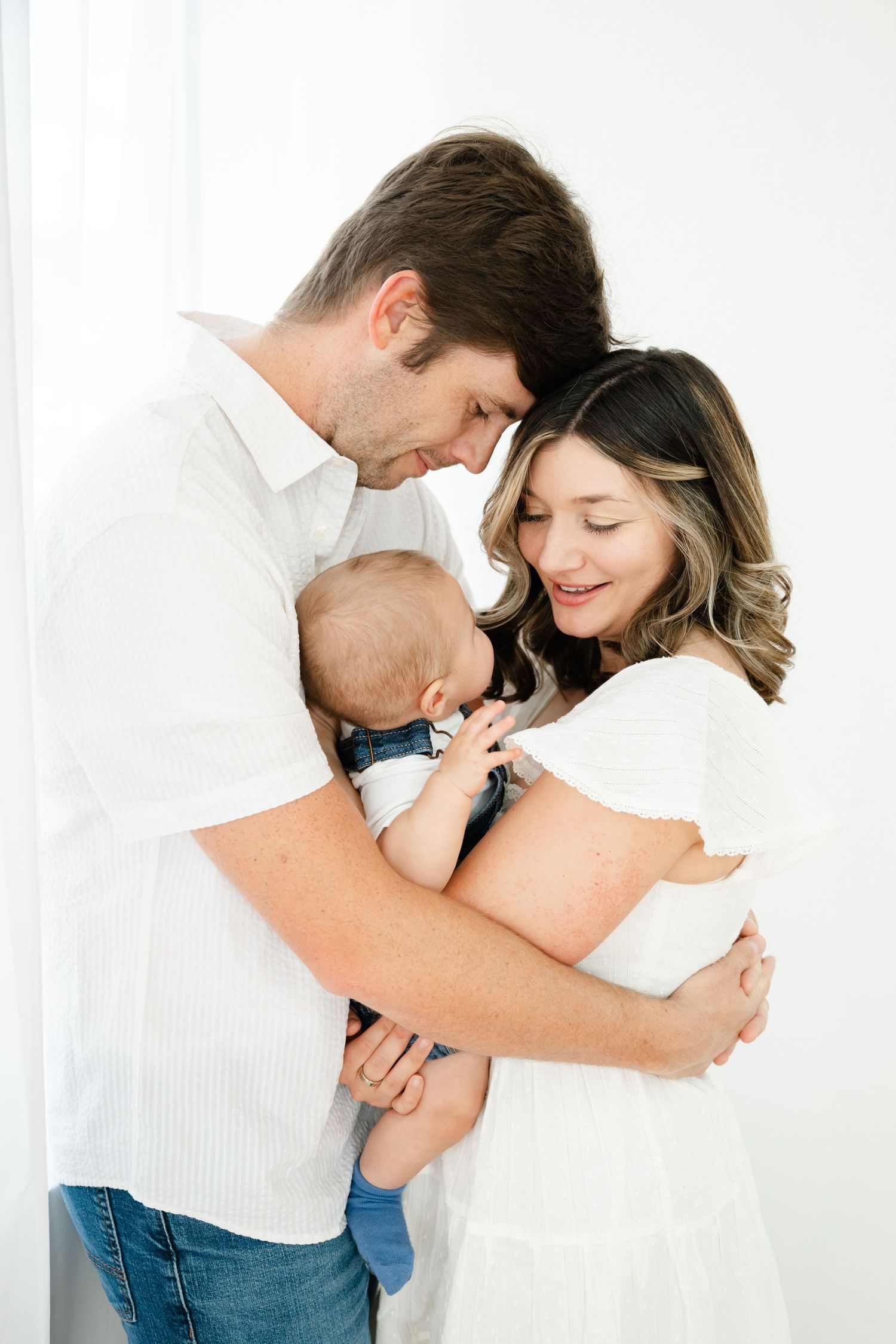 A couple in white clothing tenderly holds their newborn baby during a bright studio family portrait session.