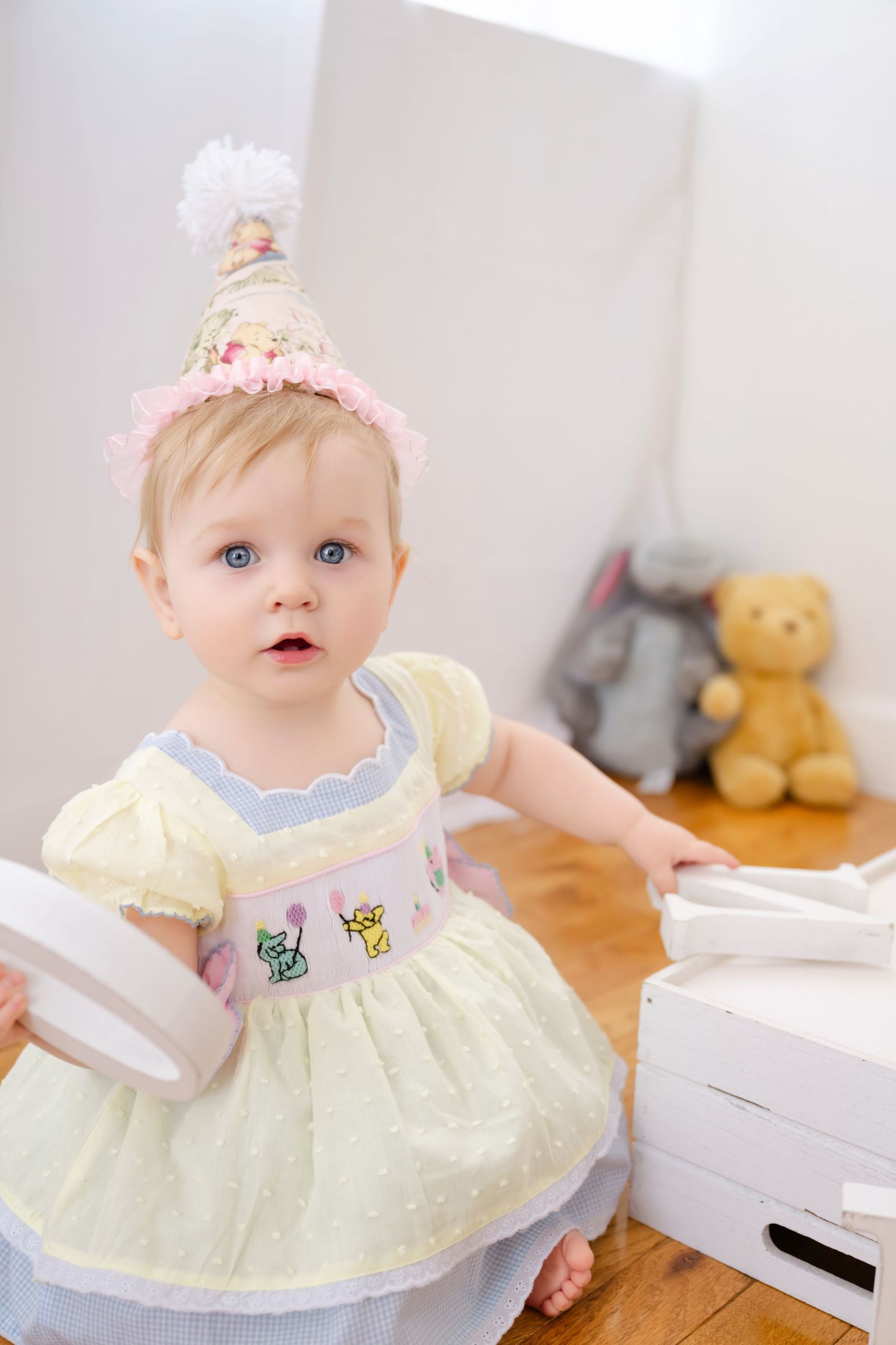 Adorable blue-eyed baby girl in yellow dress and pink party hat holding a gift box lid on her first birthday.