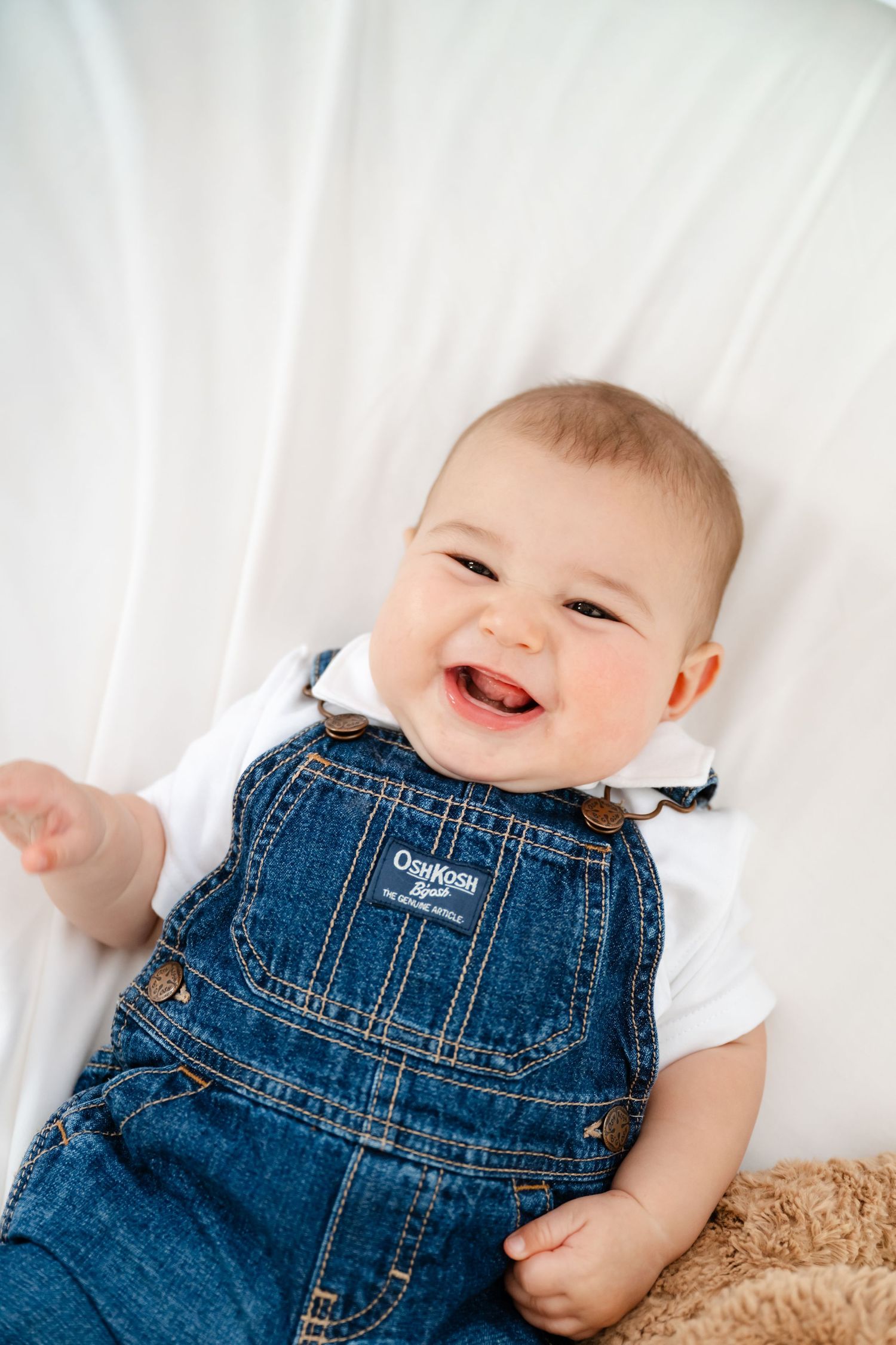 Smiling baby in OshKosh denim overalls and white onesie lying on a white background.