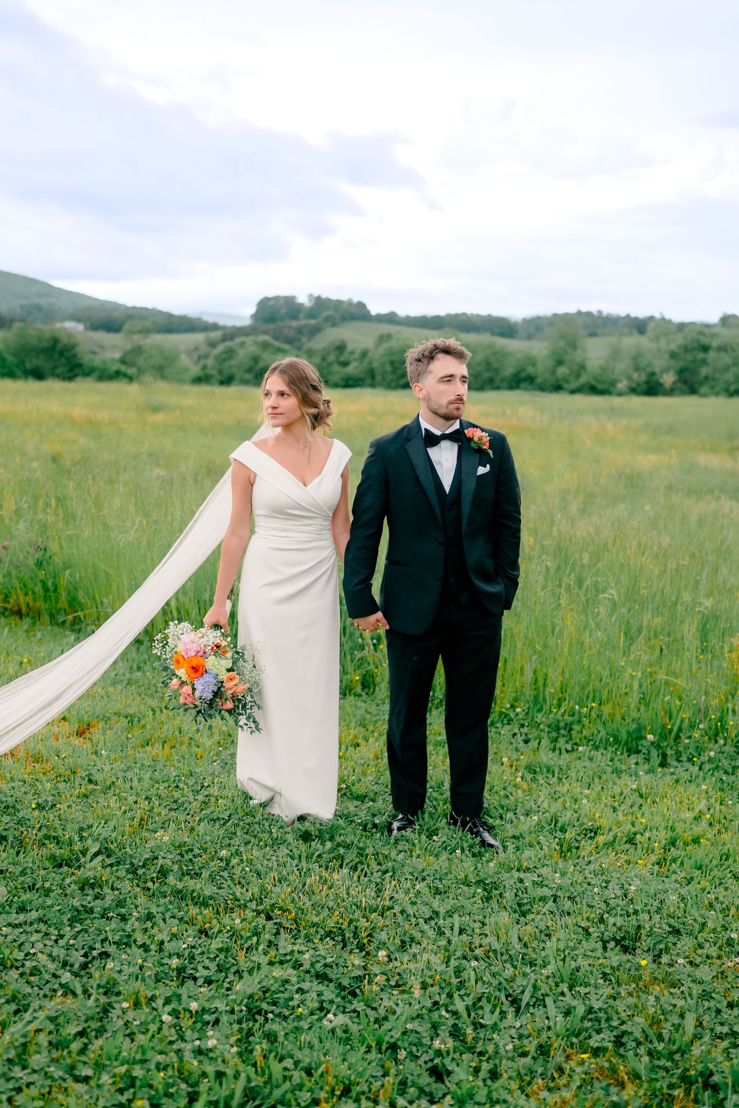 A wedding couple stands in a lush green meadow with rolling hills in the background.