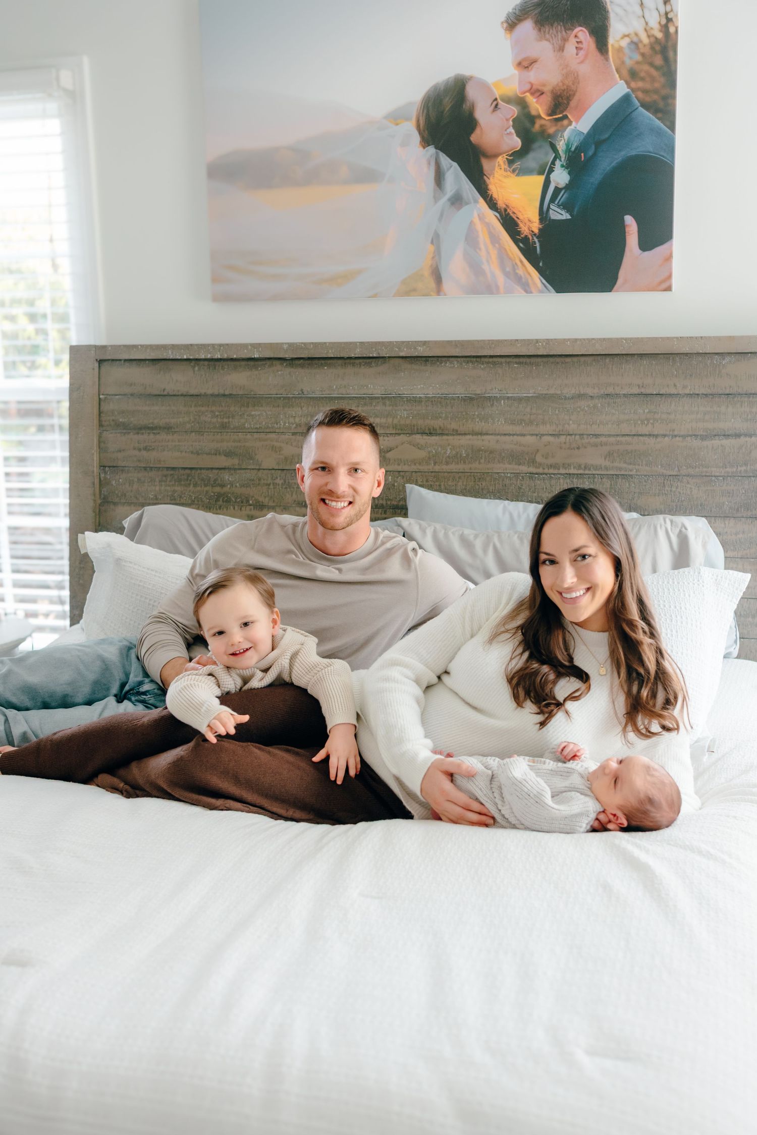 Family relaxes together on white bedding in bright bedroom.