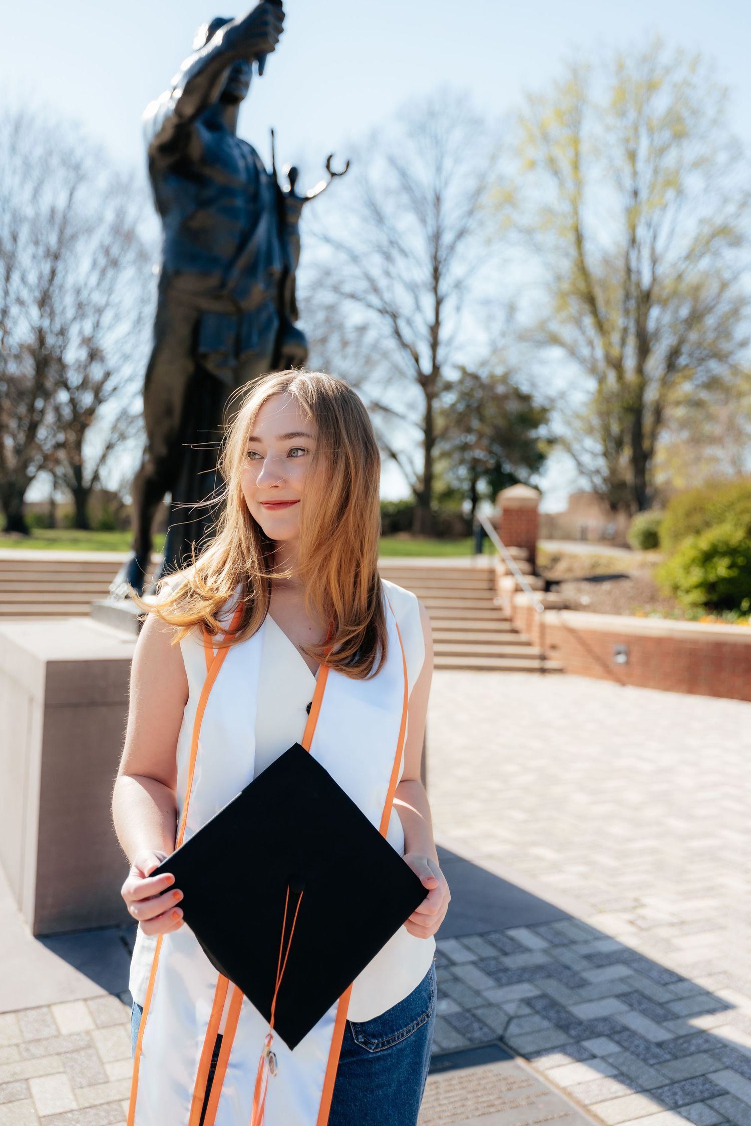 Graduate in cap and gown poses with diploma on sunny campus steps.