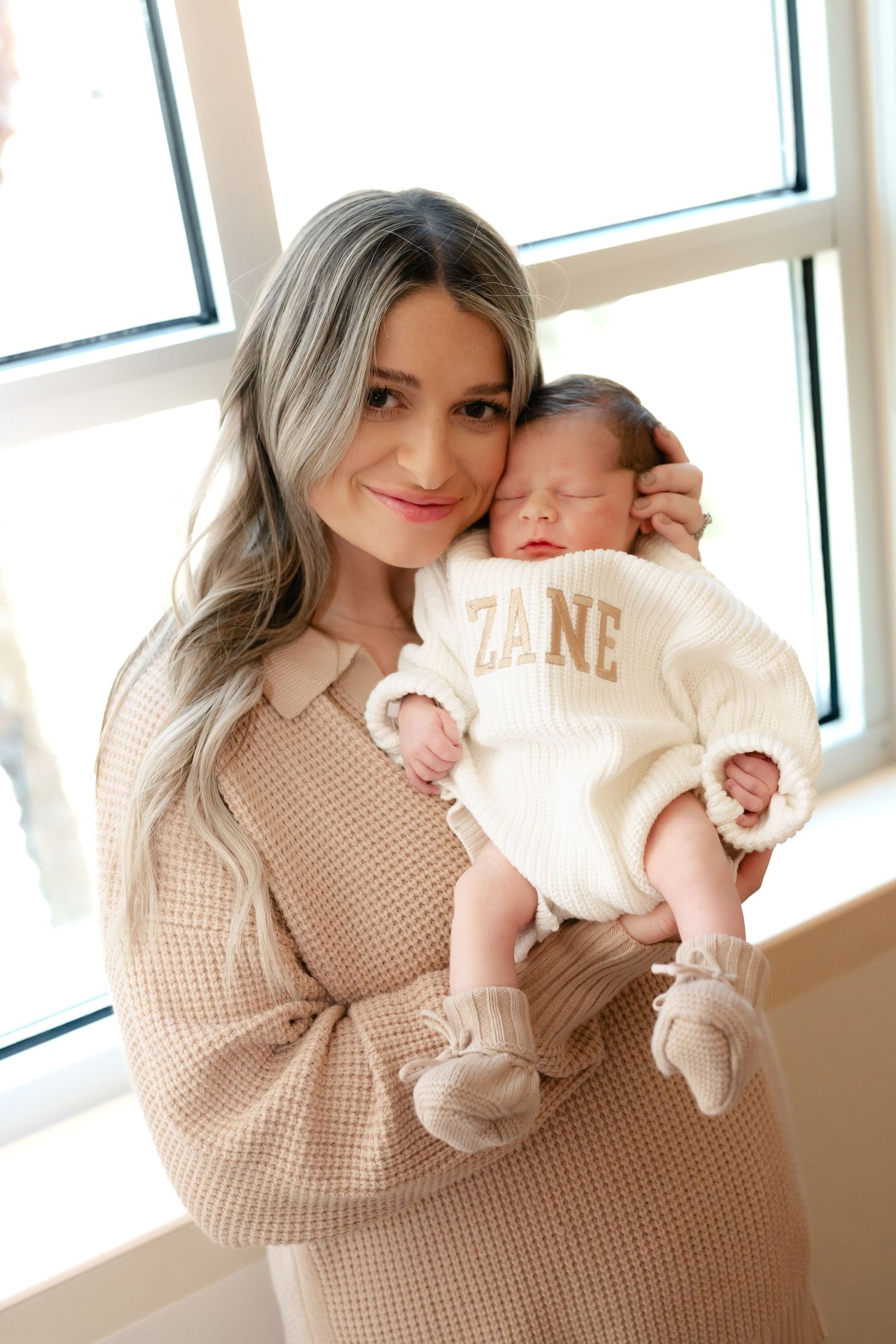 A parent in a beige sweater holds a sleeping baby wearing a cream-colored outfit with 'ZANE' text by a bright window.