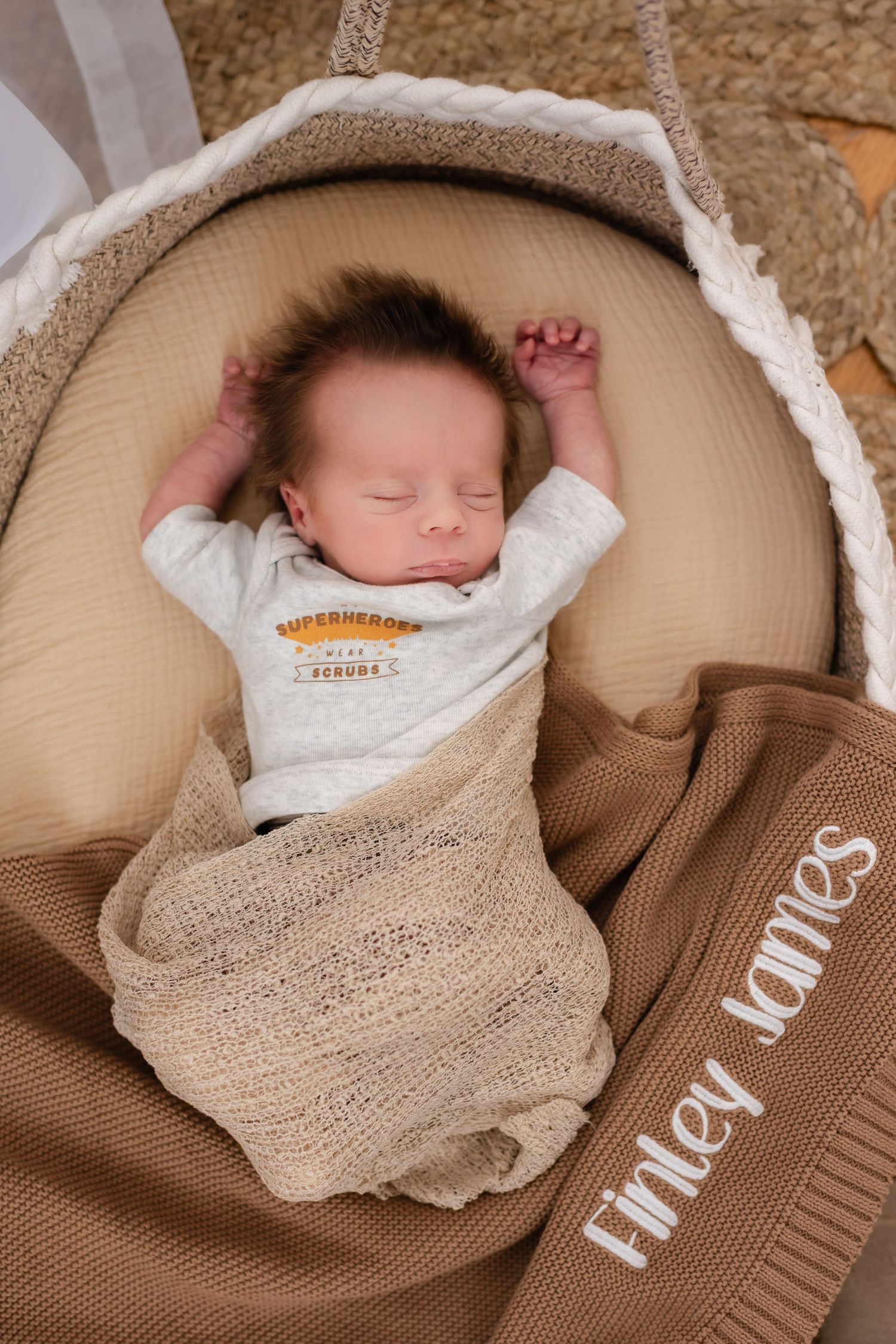 Sleeping infant wrapped in beige blanket rests peacefully in tan-colored baby lounger with Tinley James logo.