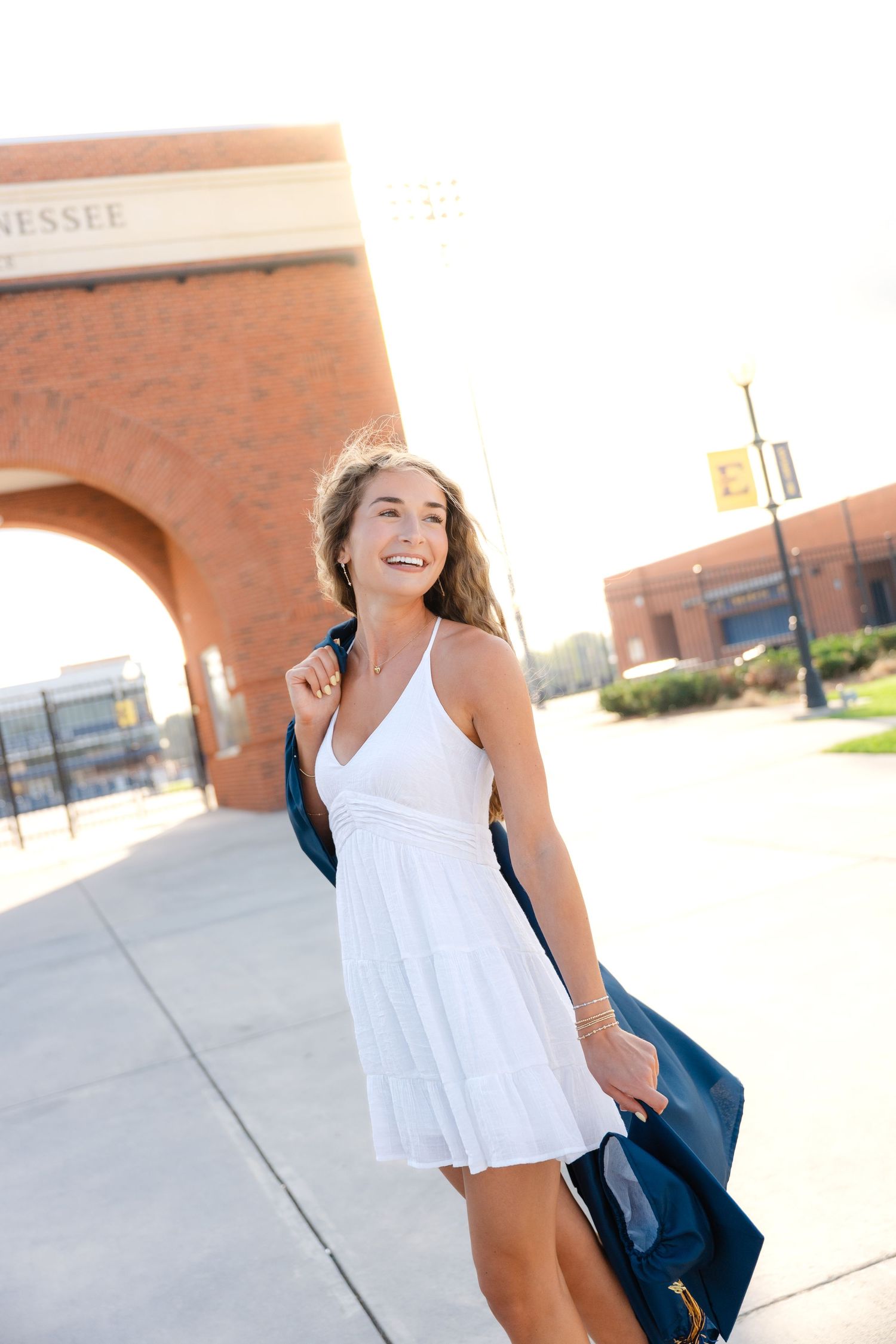 Smiling young woman in white dress holding graduation gown outside Tennessee university brick arch on sunny day.