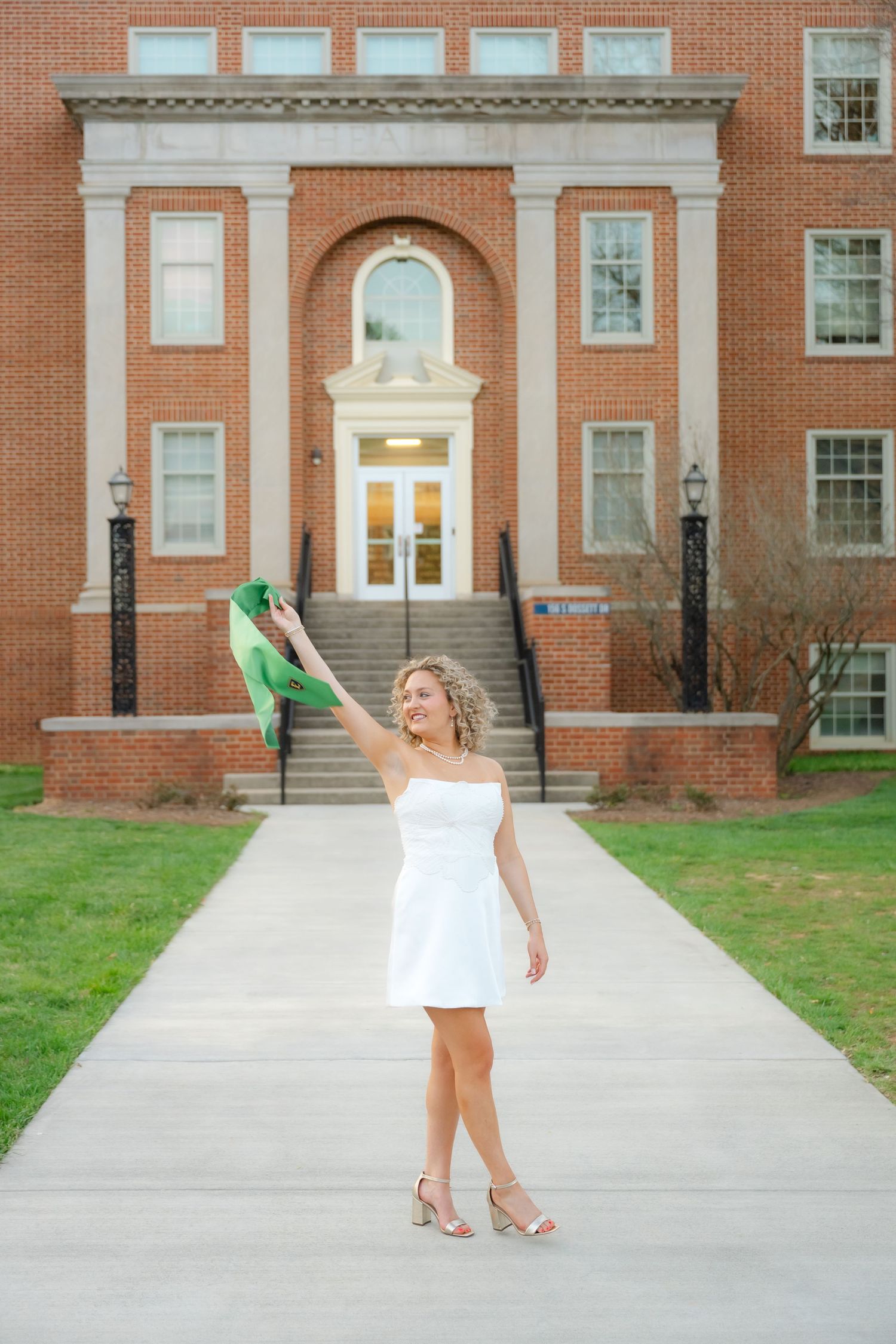Young woman in white dress holding green heels, celebrating outdoors in front of a classic brick building.