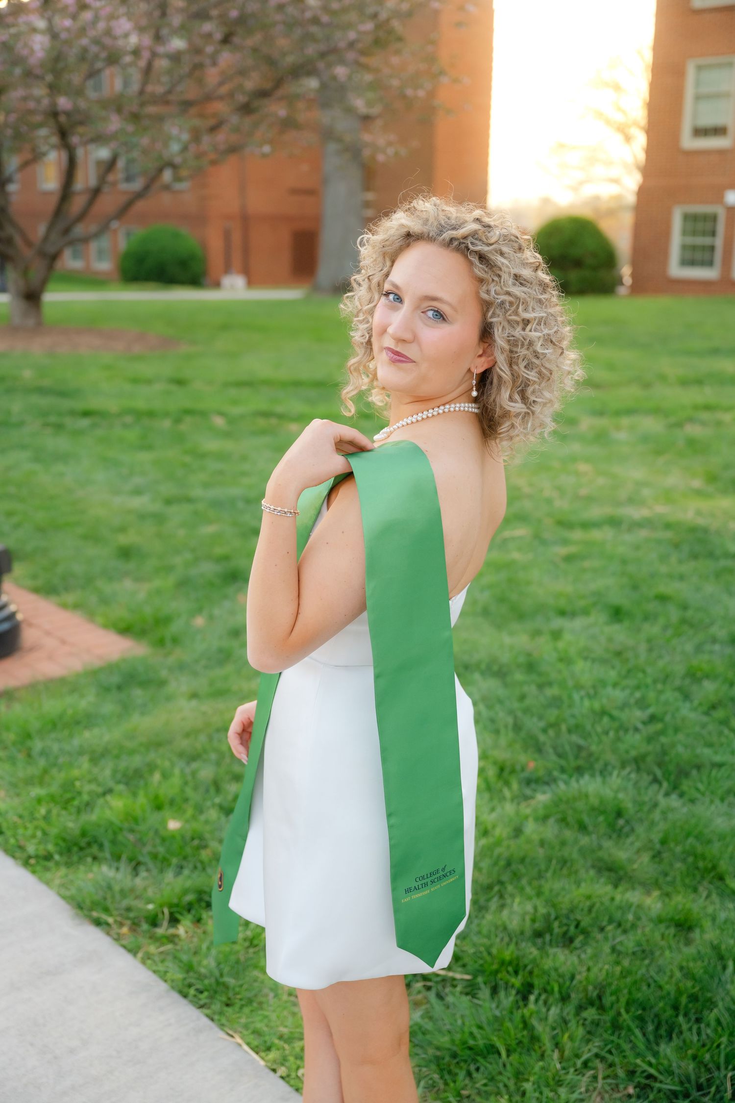 Young woman in white dress holding green graduation stole poses outdoors on college campus at golden hour.
