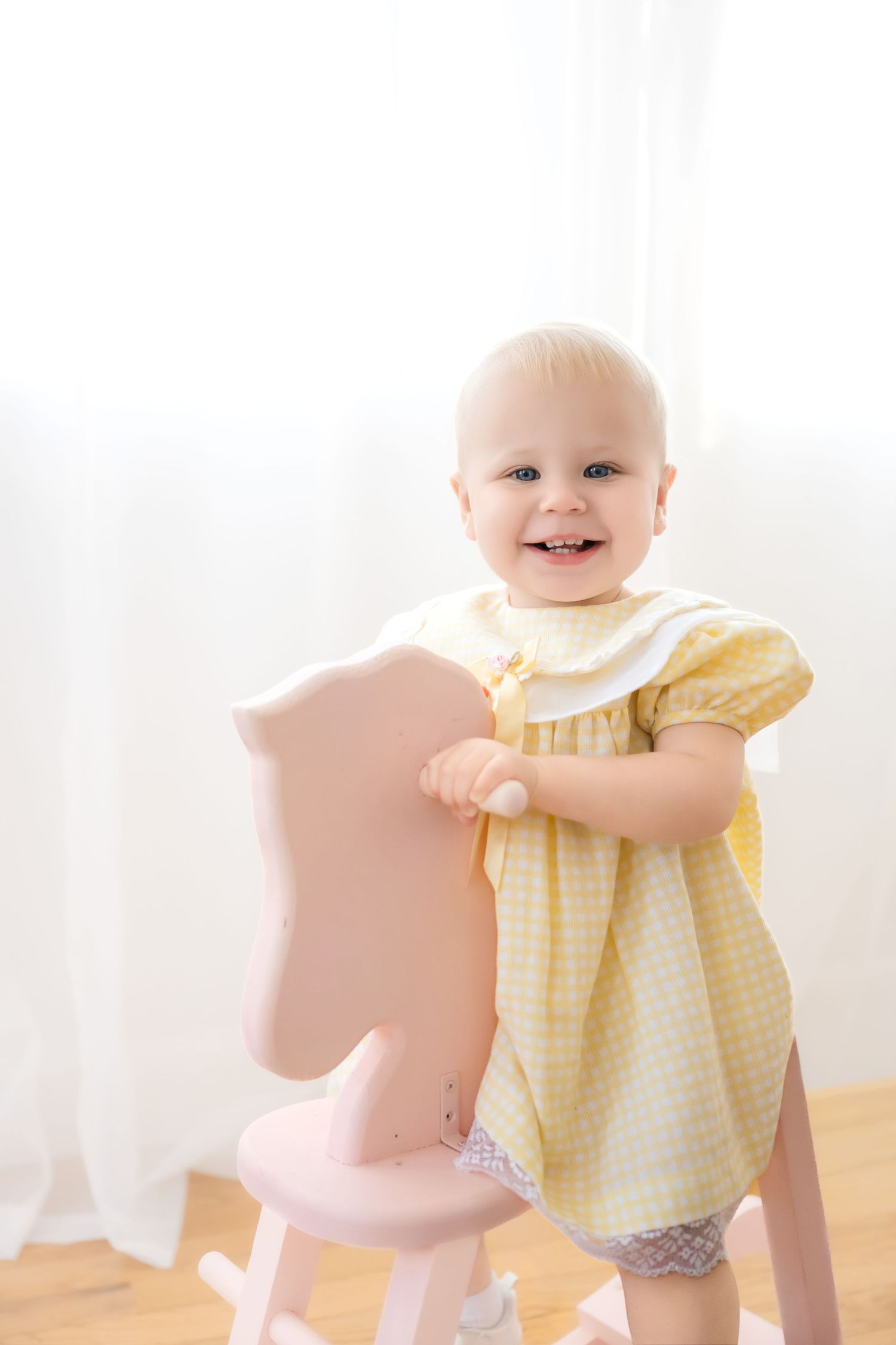 Smiling baby girl in yellow gingham dress standing beside a pink rocking horse toy in a bright white room.