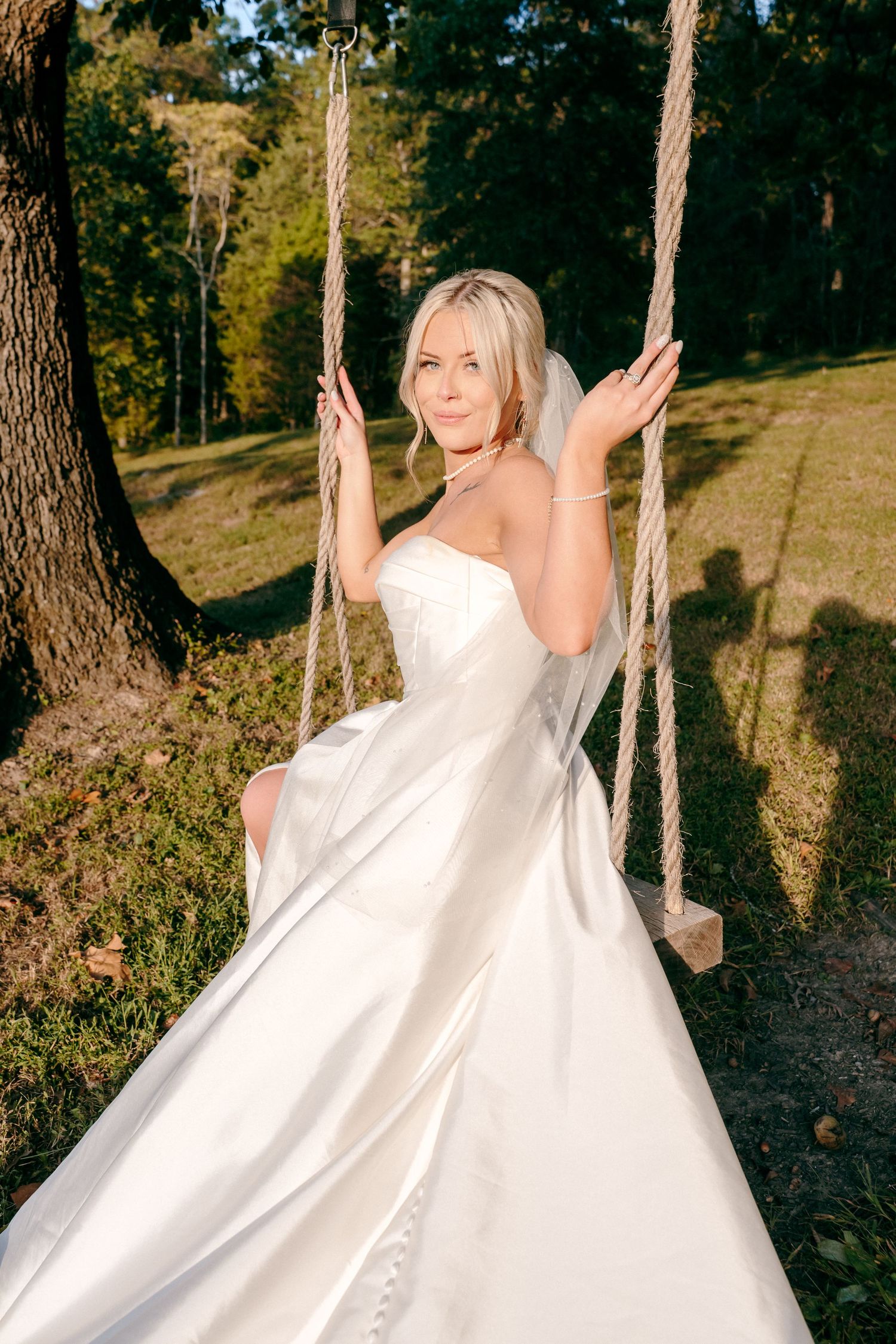 Bride in flowing white gown poses gracefully on a rustic rope swing in a wooded setting.