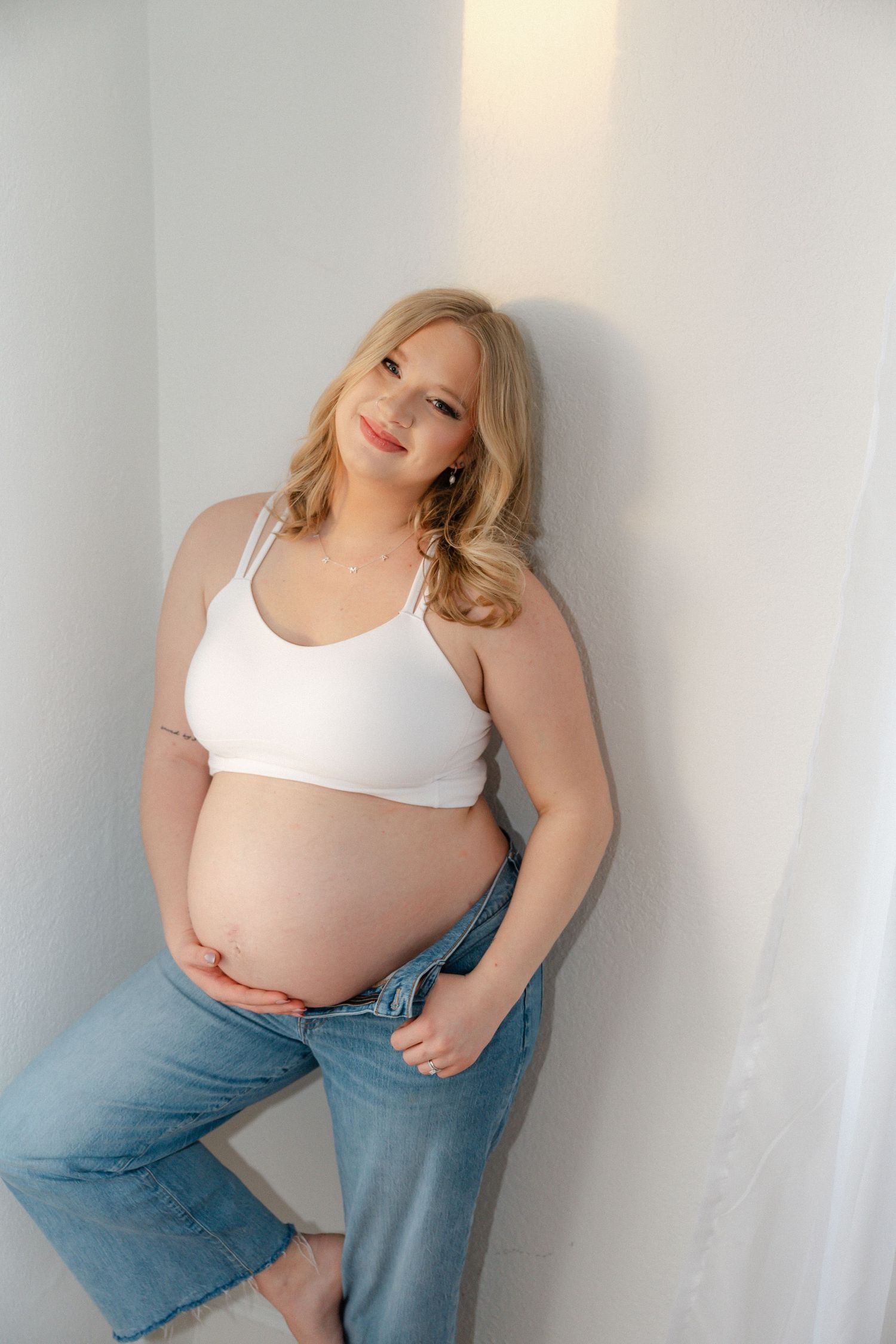 Expectant mother in white crop top and jeans poses against a white wall showing her pregnant belly.