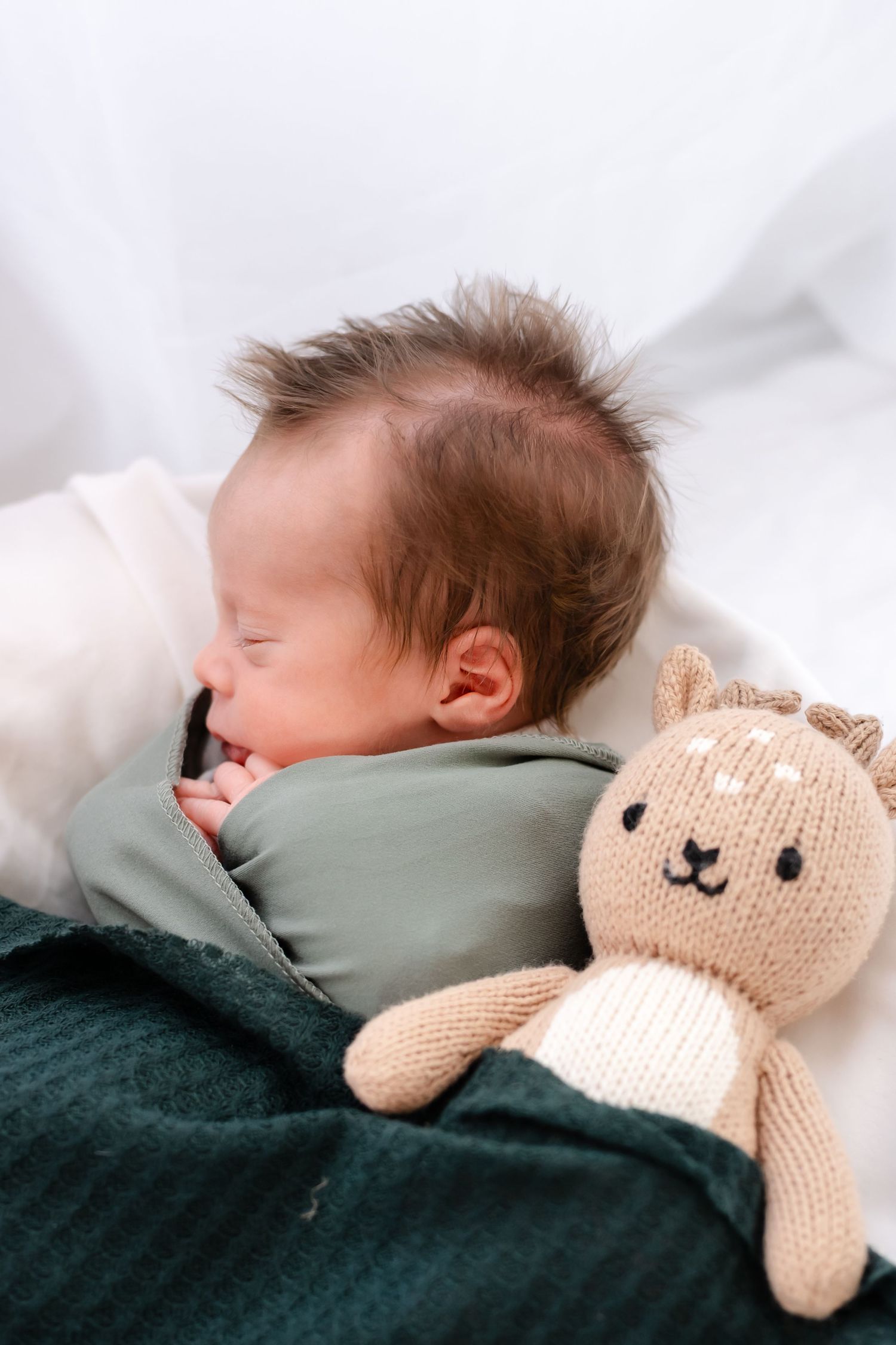 A sleeping baby lies peacefully in bed next to a knitted beige teddy bear on dark green bedding.