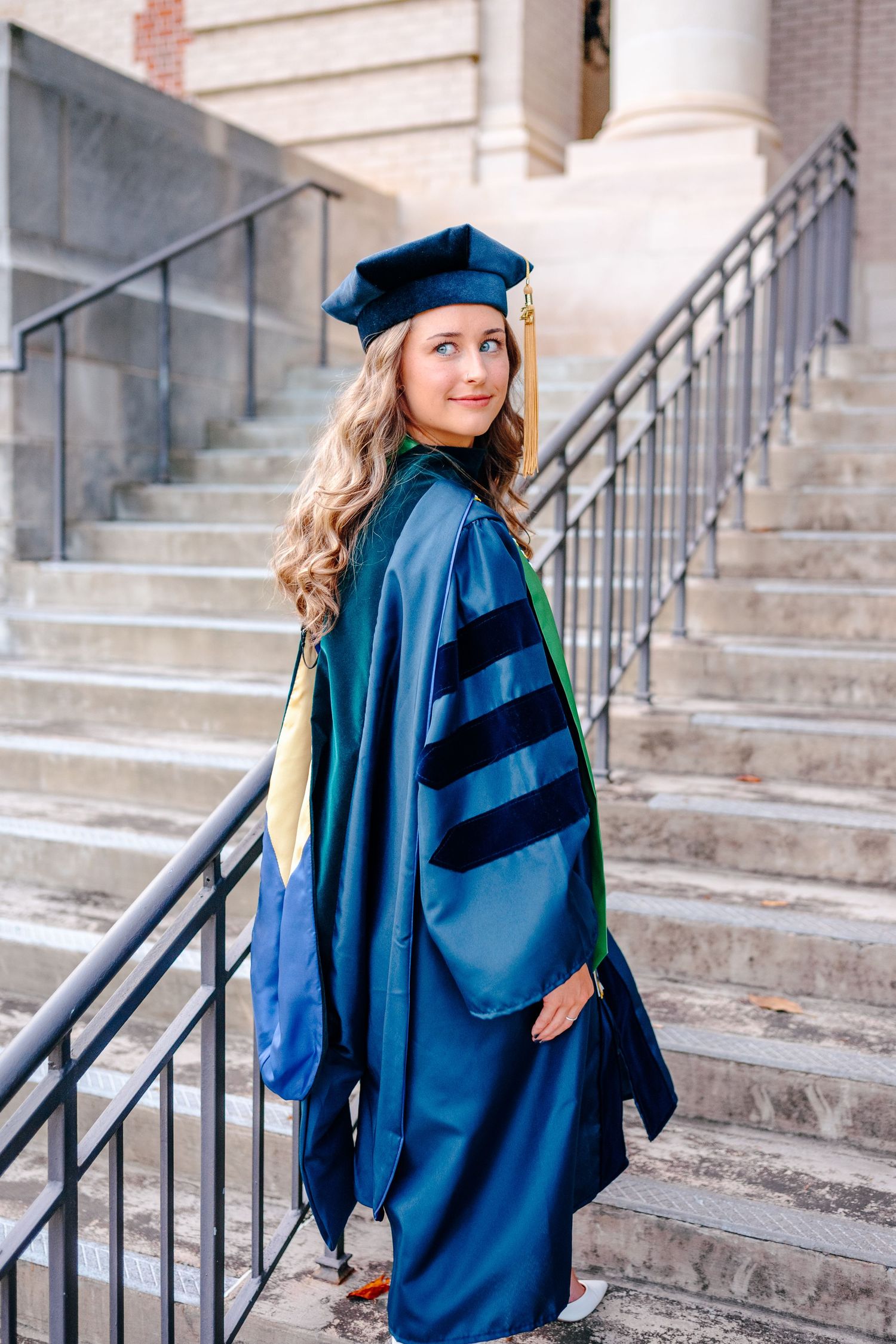 Graduate in blue academic regalia poses on stone steps with metal railing.