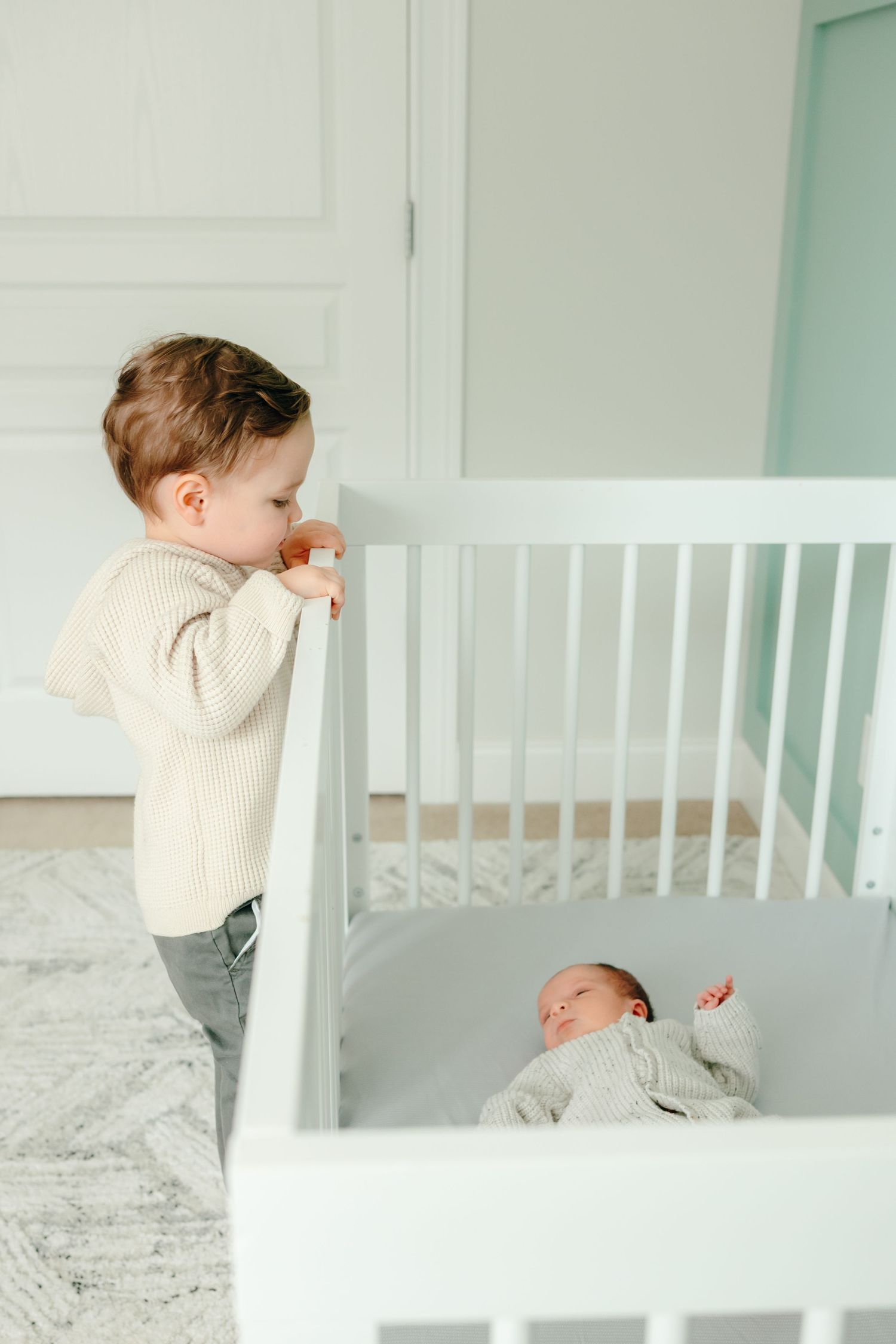 Toddler peeks through white crib bars at baby sibling.