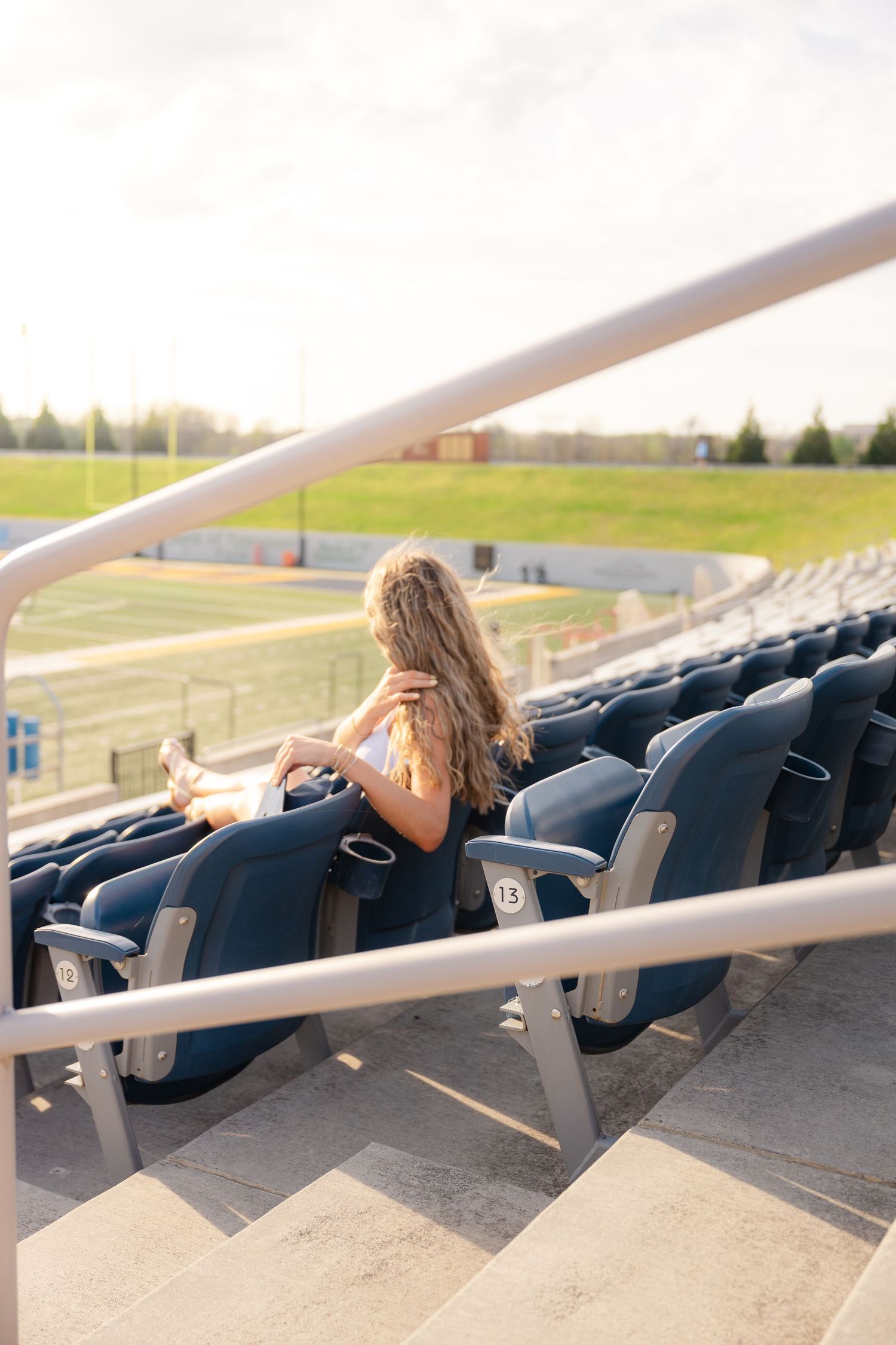 Young girl with long hair sitting alone in empty stadium bleachers, overlooking a green sports field on a sunny day.