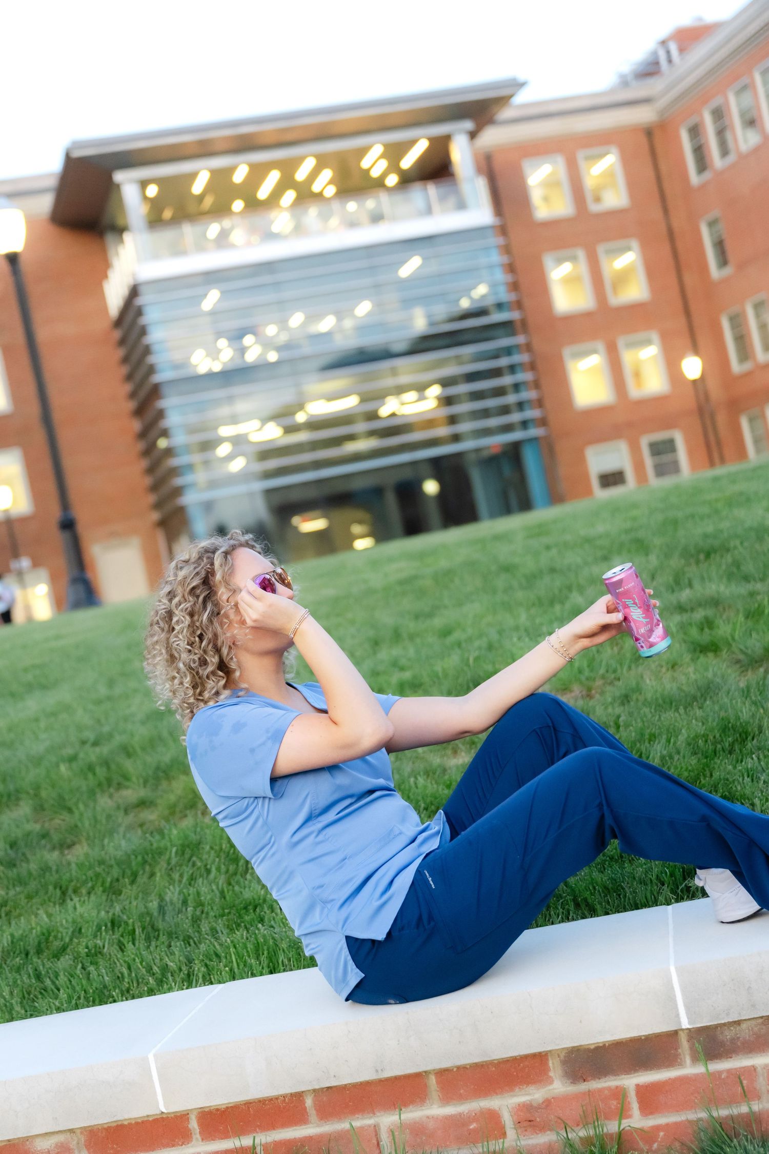 Young woman with curly hair relaxing on a brick ledge outdoors, holding a pink can near a modern university building.