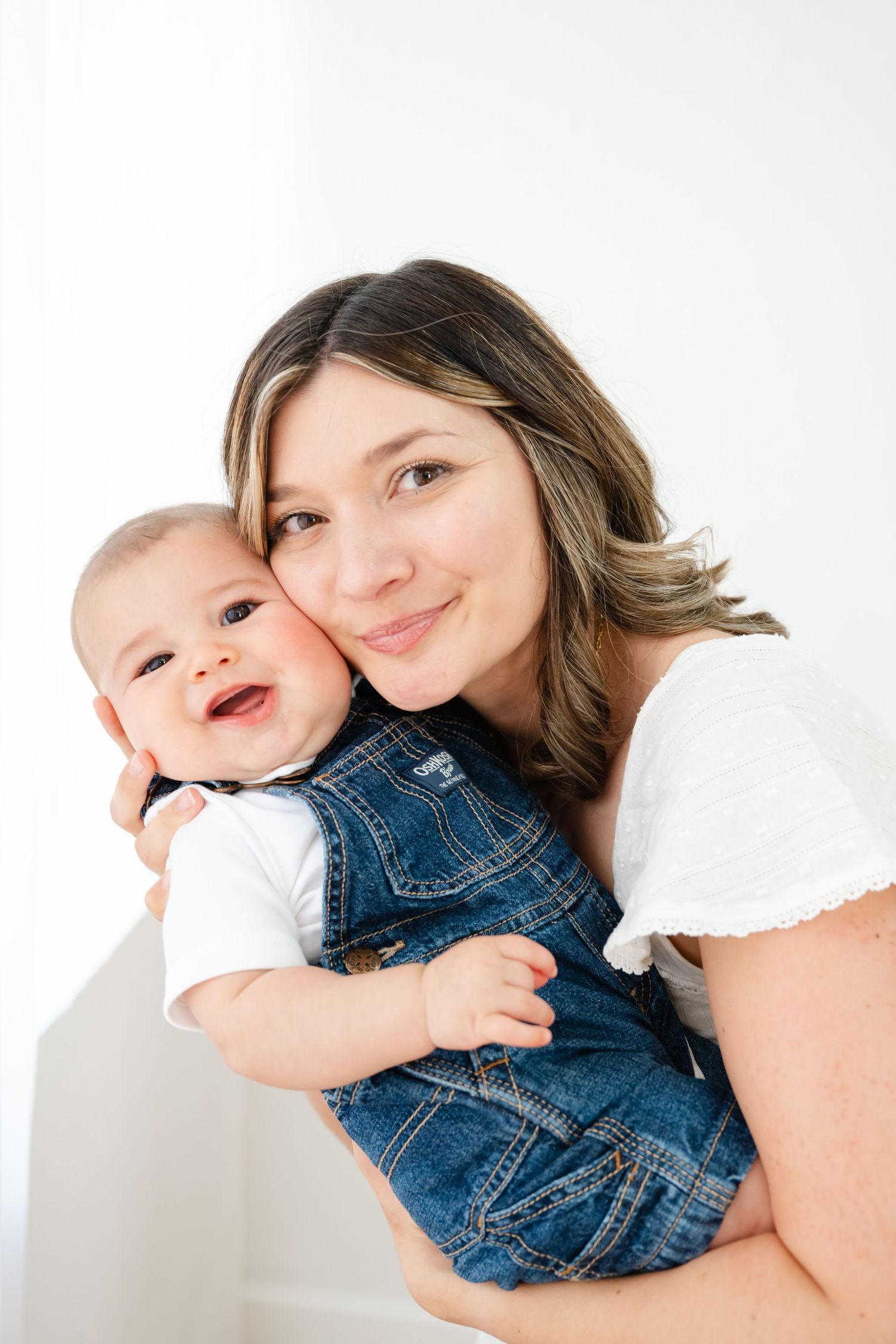 A smiling mother holds her happy baby dressed in denim overalls against a white background.