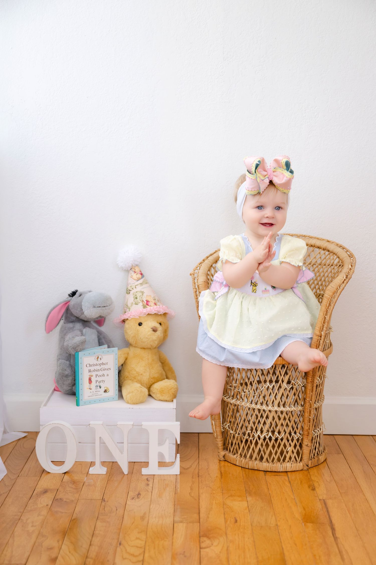 Baby girl in yellow dress sits in wicker chair beside Winnie the Pooh toys and ONE sign for first birthday.
