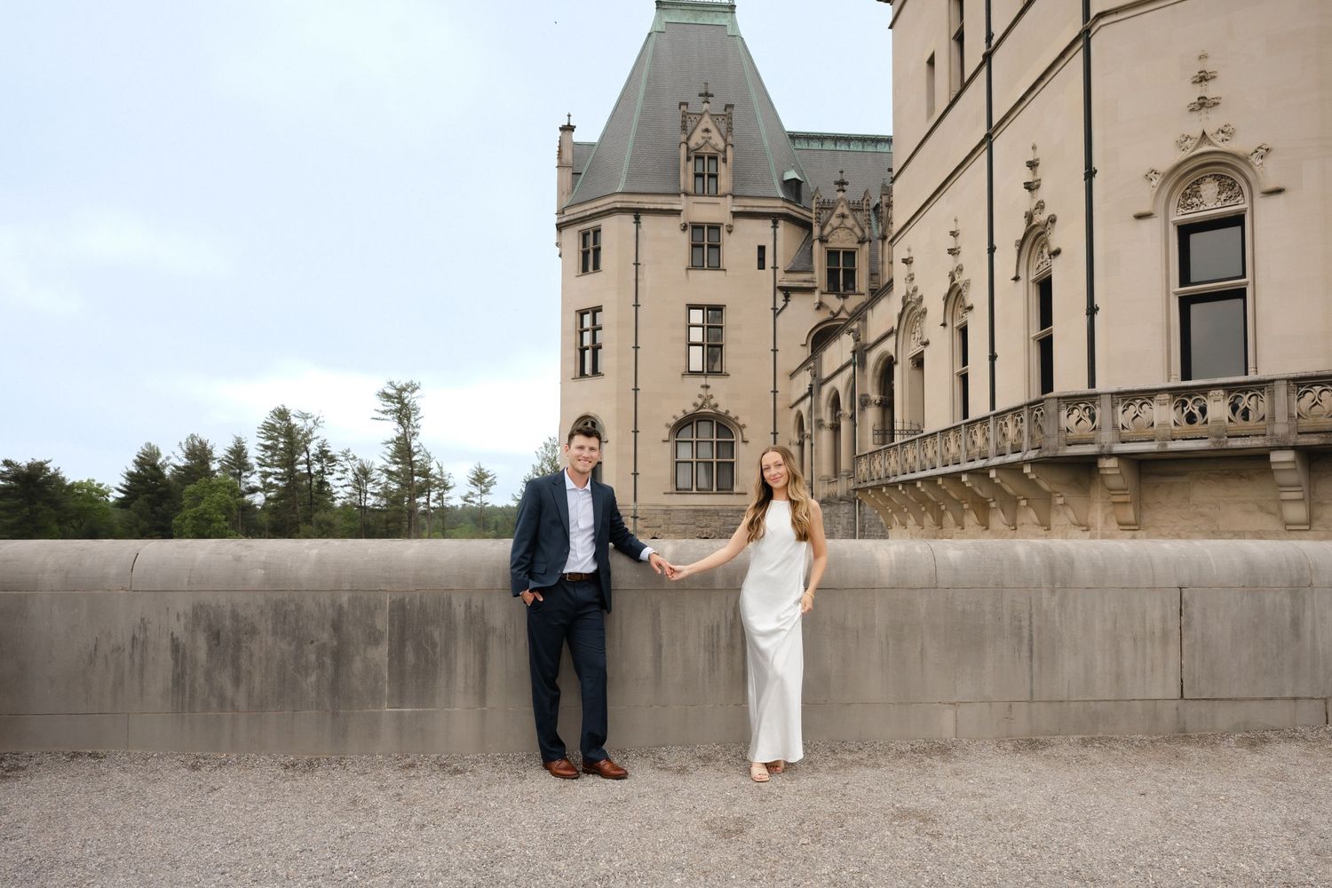 Couple holding hands in front of Biltmore Estate castle, woman in white dress, man in navy suit, outdoor engagement photo.
