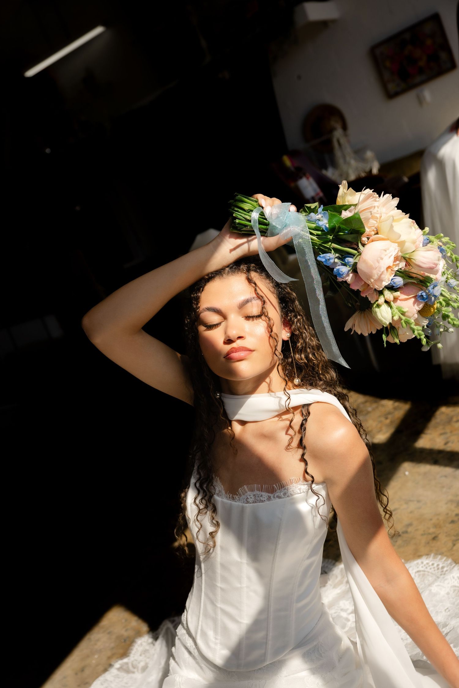 Bride in white corset gown holds floral bouquet overhead with eyes closed, bathed in sunlight indoors.
