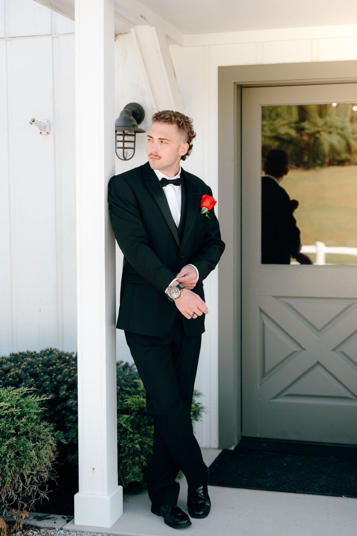 A young ring bearer in a black tuxedo poses playfully on a white porch.