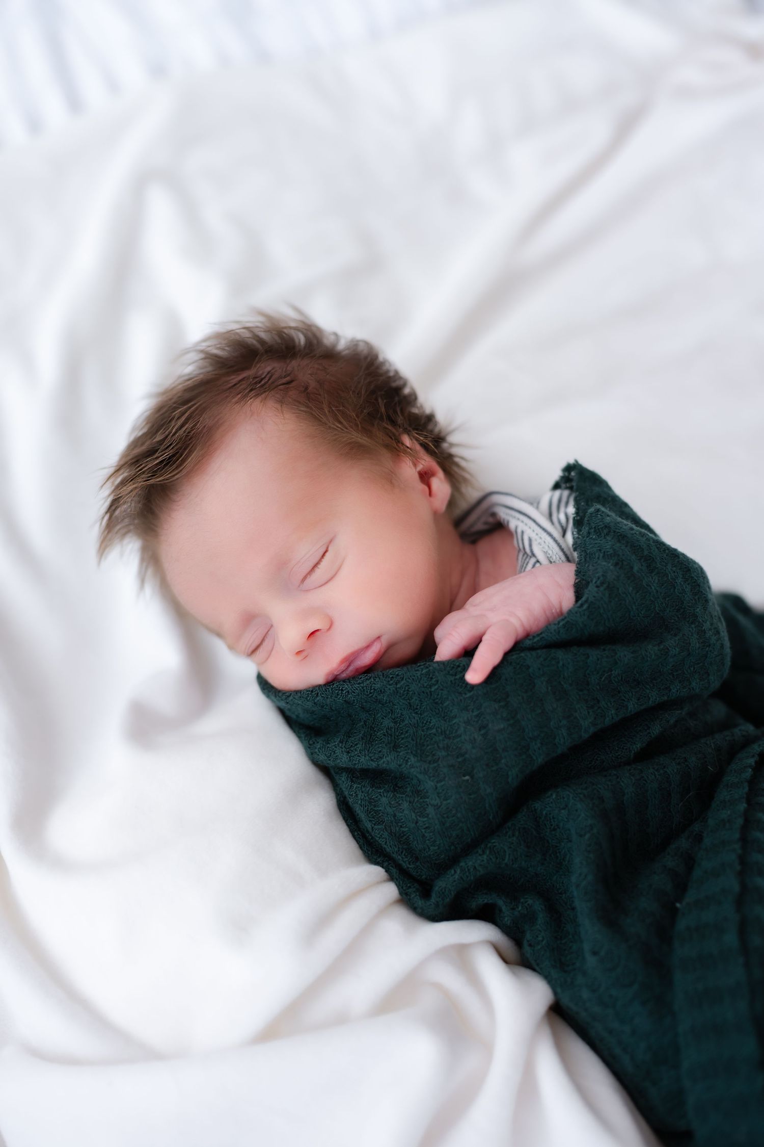 A sleeping baby wrapped in a dark blanket rests peacefully on white bedding.