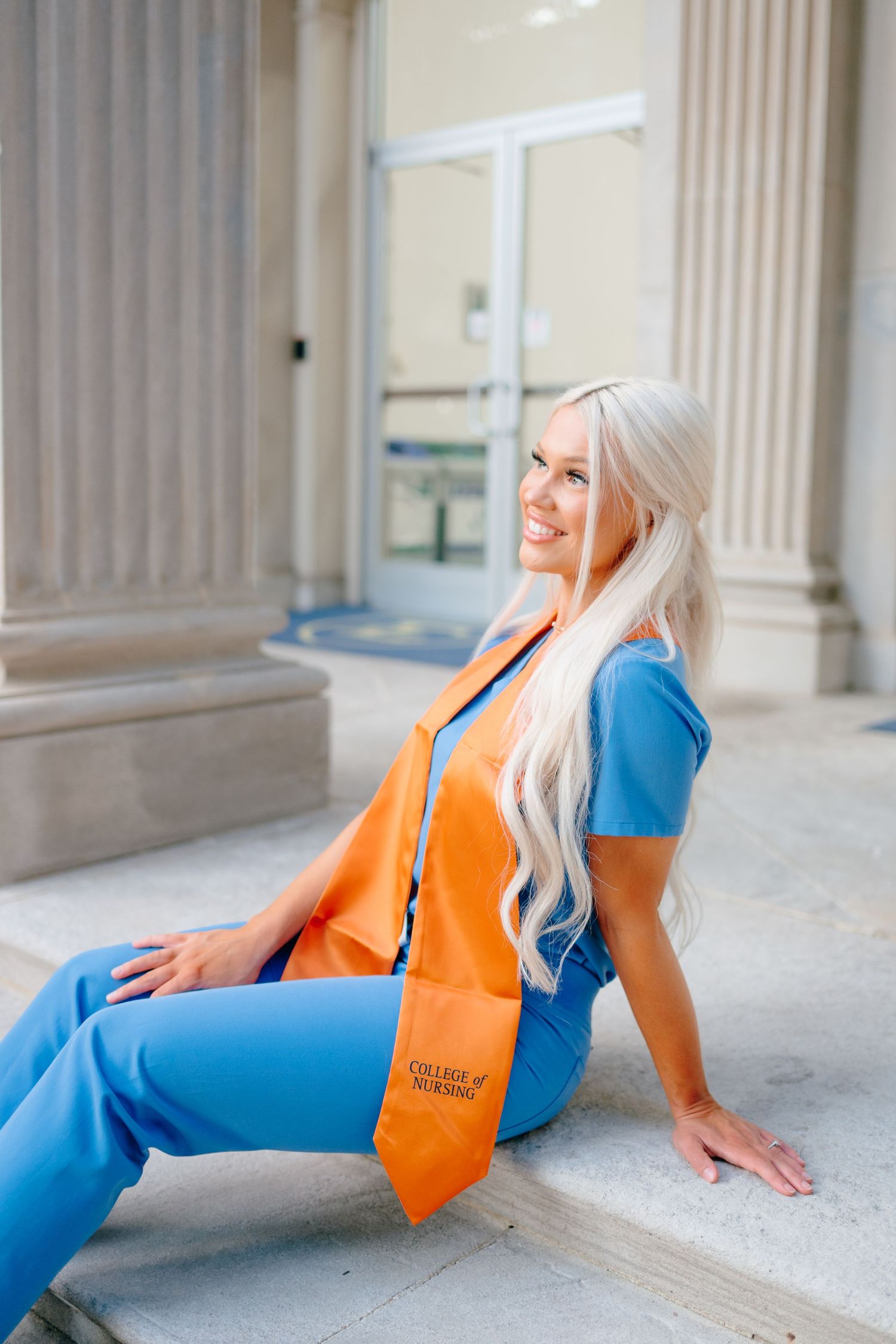 Graduate in blue outfit and orange stole poses on campus steps.