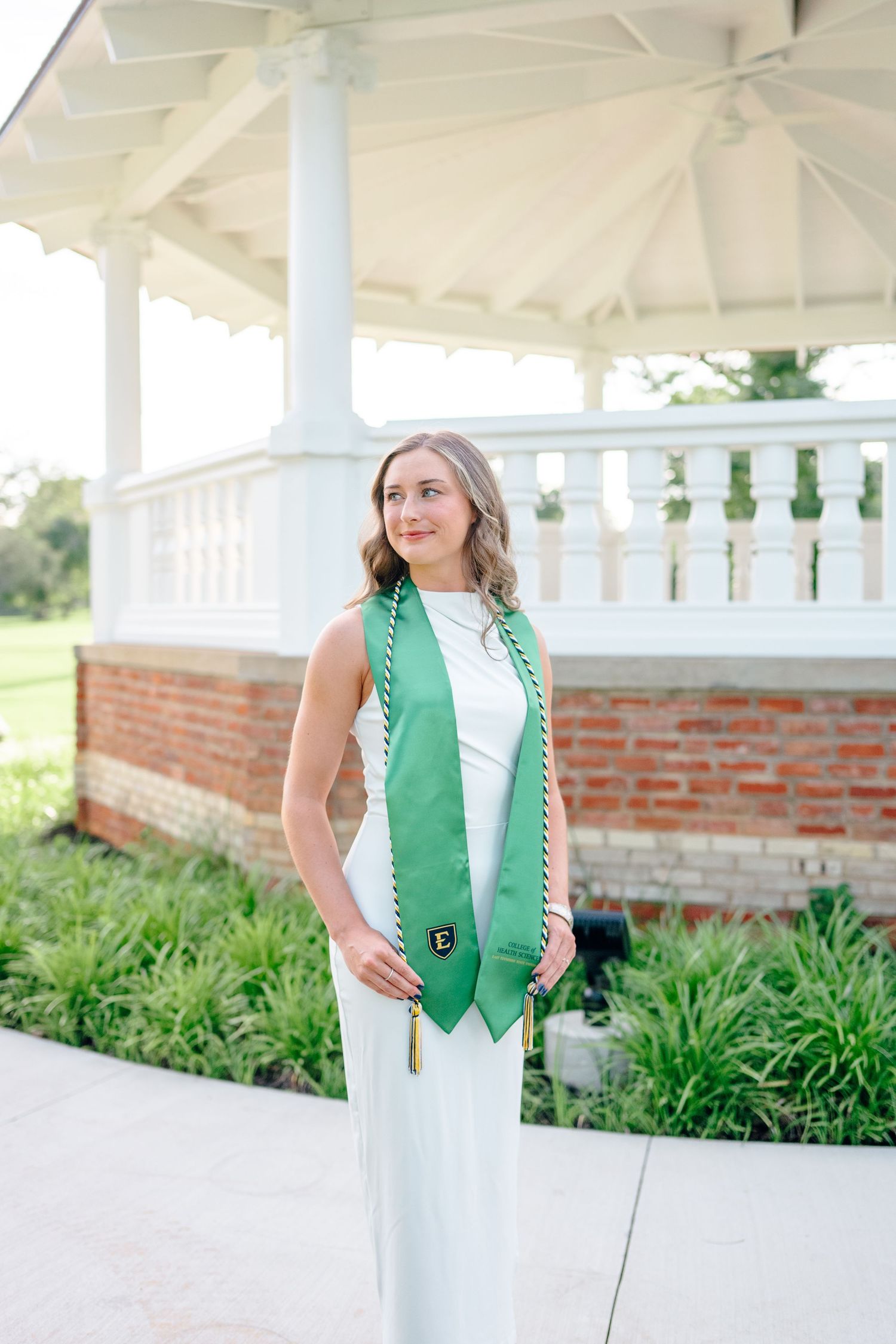 Graduate wearing a green vest and white dress poses under a white gazebo.