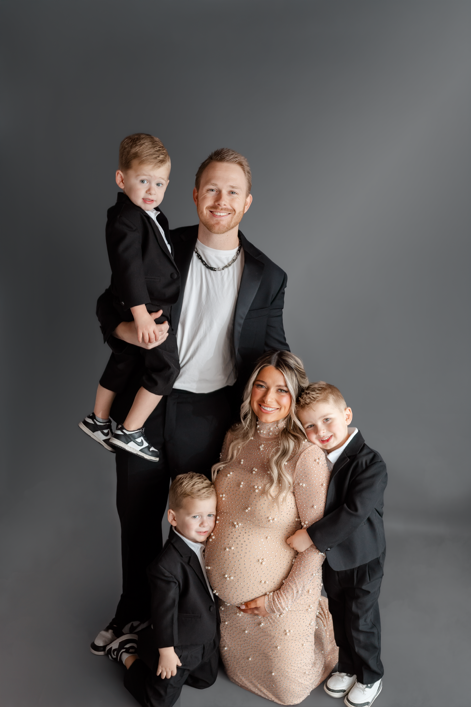 A family maternity photo shoot featuring a pregnant woman in a beige dress with two young boys in black suits.