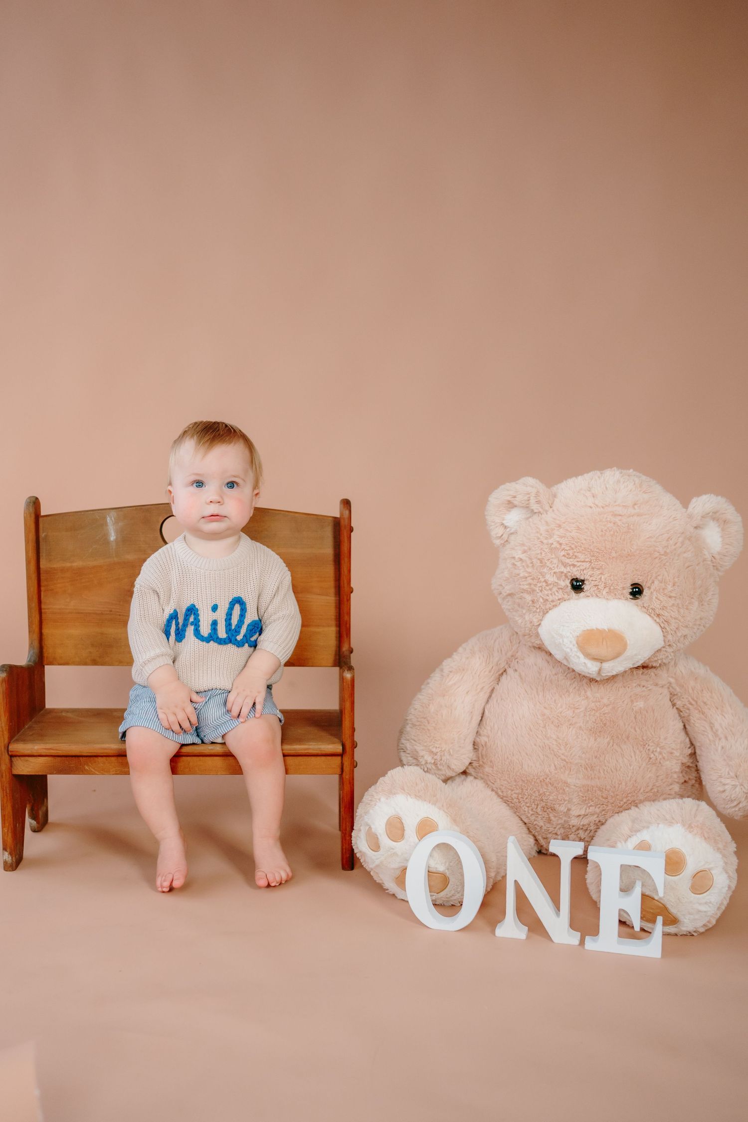 Baby sitting in wooden chair with teddy bear and 'ONE' letters for first birthday photo.