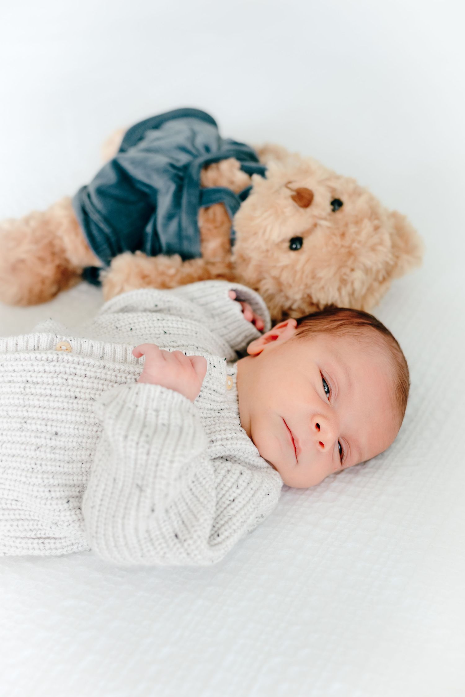 Baby in white knit sweater lying next to plush teddy bear on white background.