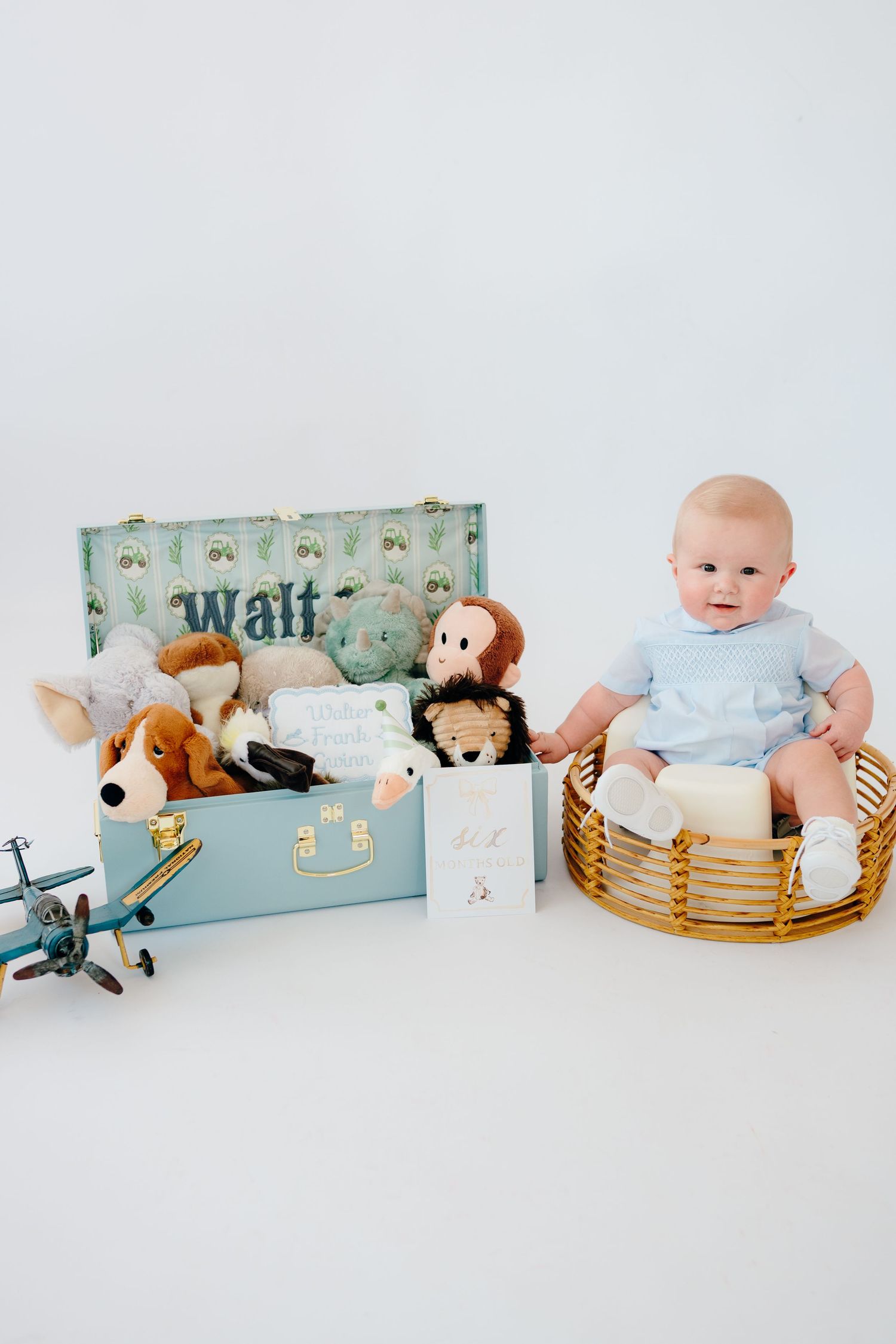 Baby with stuffed animals and toys arranged in vintage suitcase and basket.