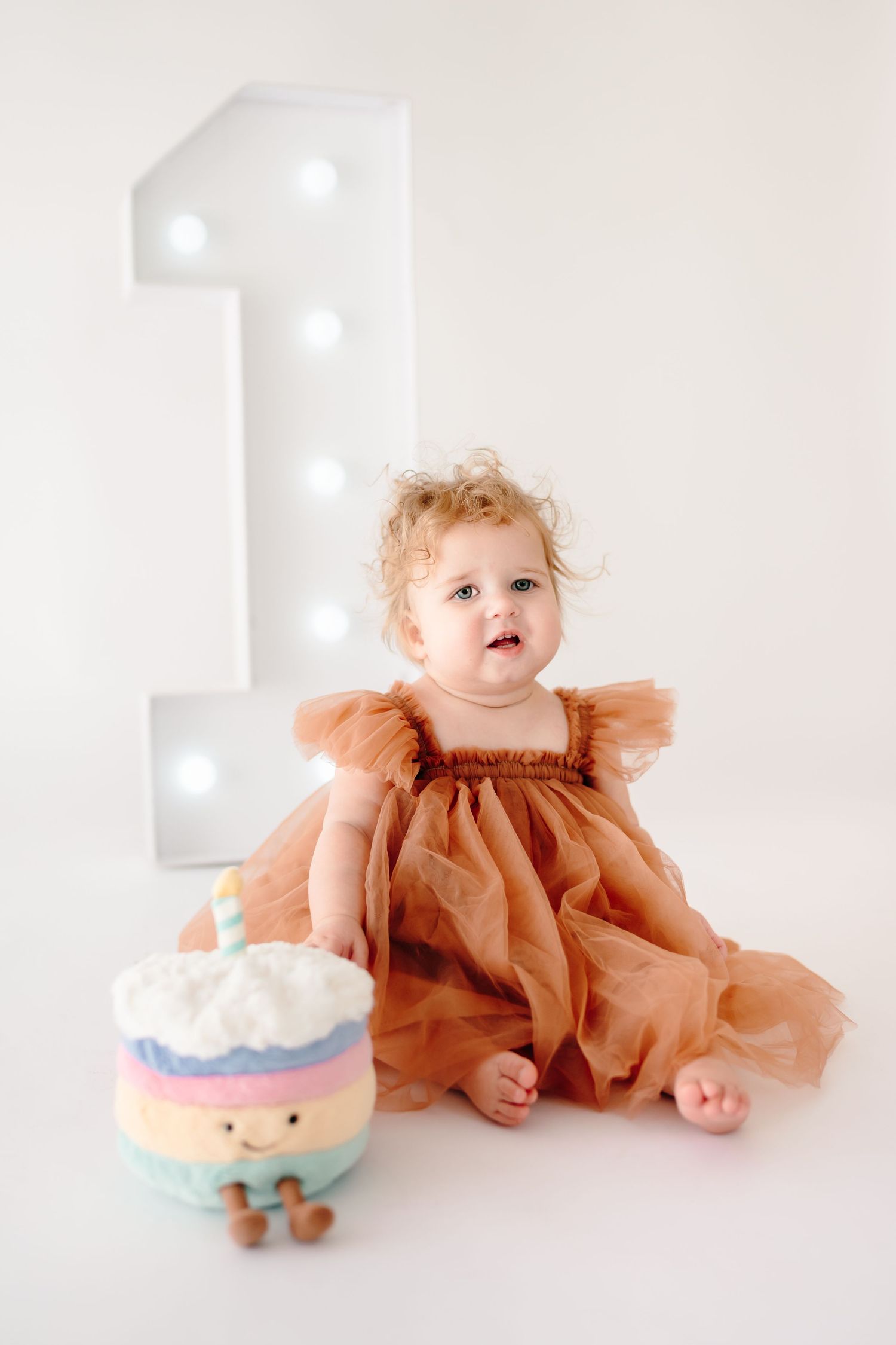 Baby in rust-colored dress sits near decorative birthday cake on white background.