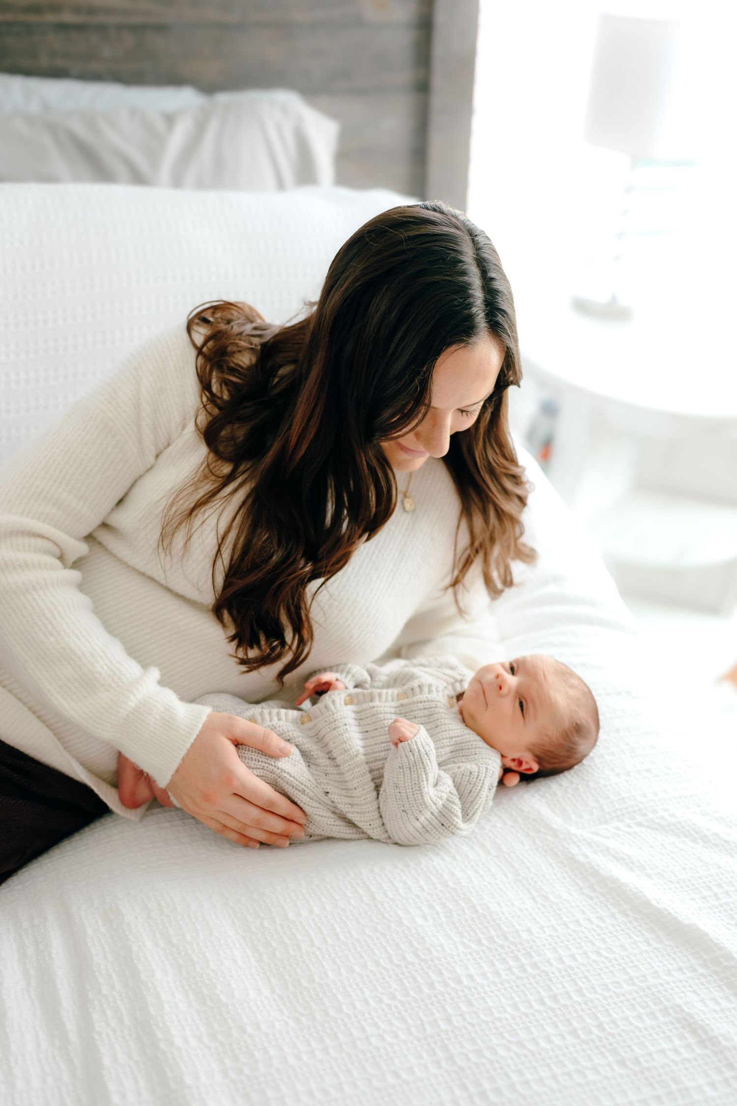 Mother in white sweater leans over baby lying on textured bedding.